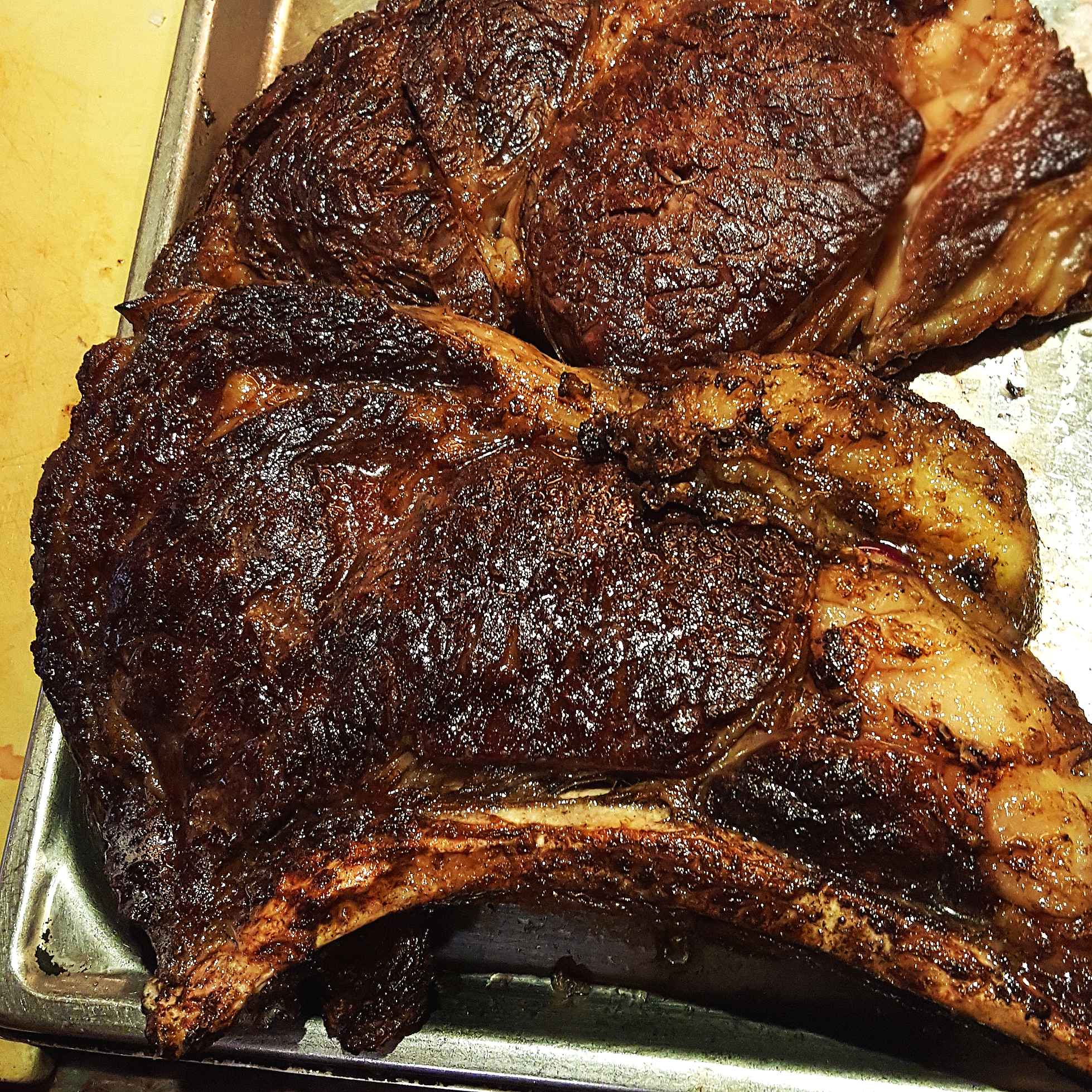 A grilled beef steak with a dark, caramelized crust, resting on a large metal tray.