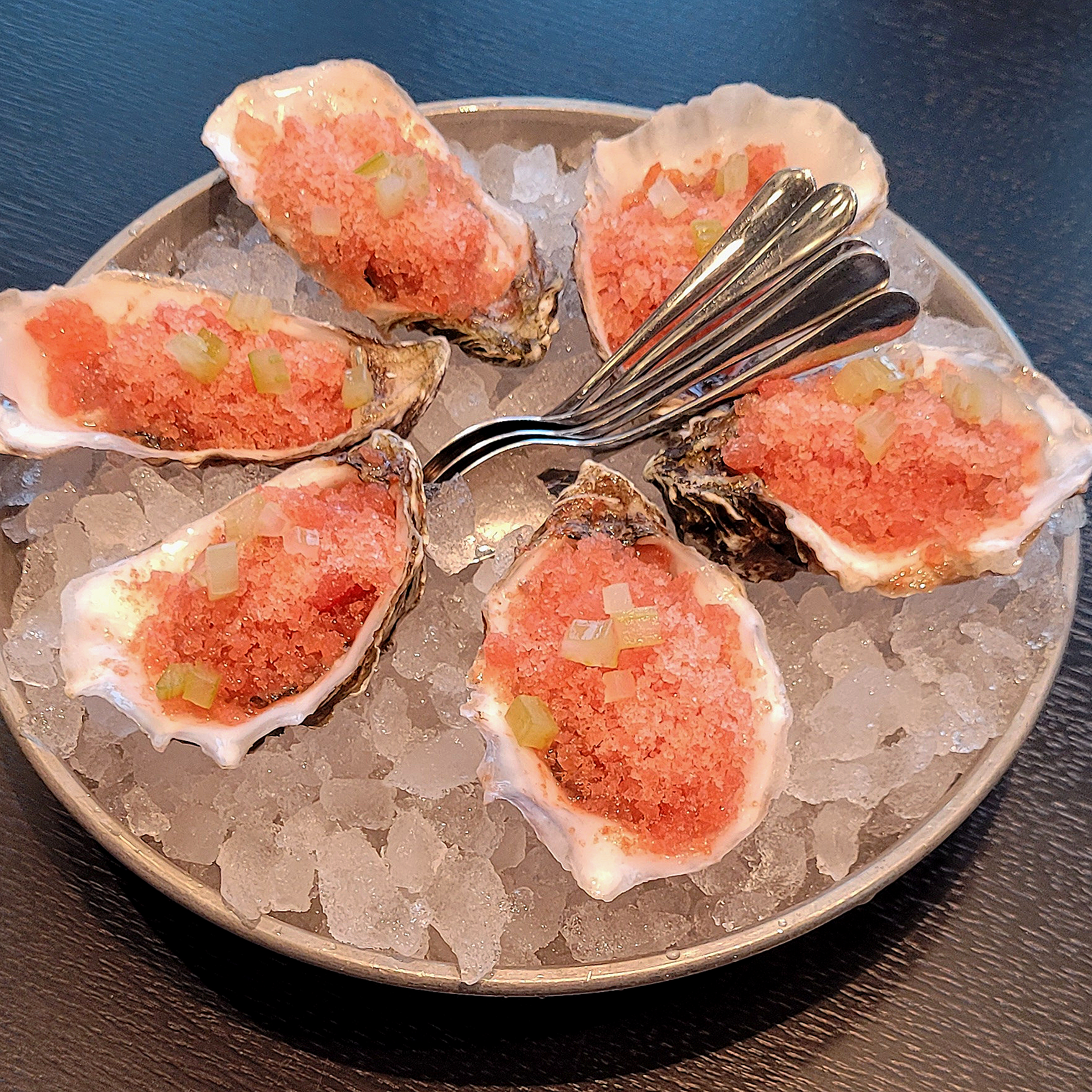 A round metal tray filled with crushed ice, topped with several oysters on the half shell, garnished with small diced vegetables, and accompanied by a few silver forks.