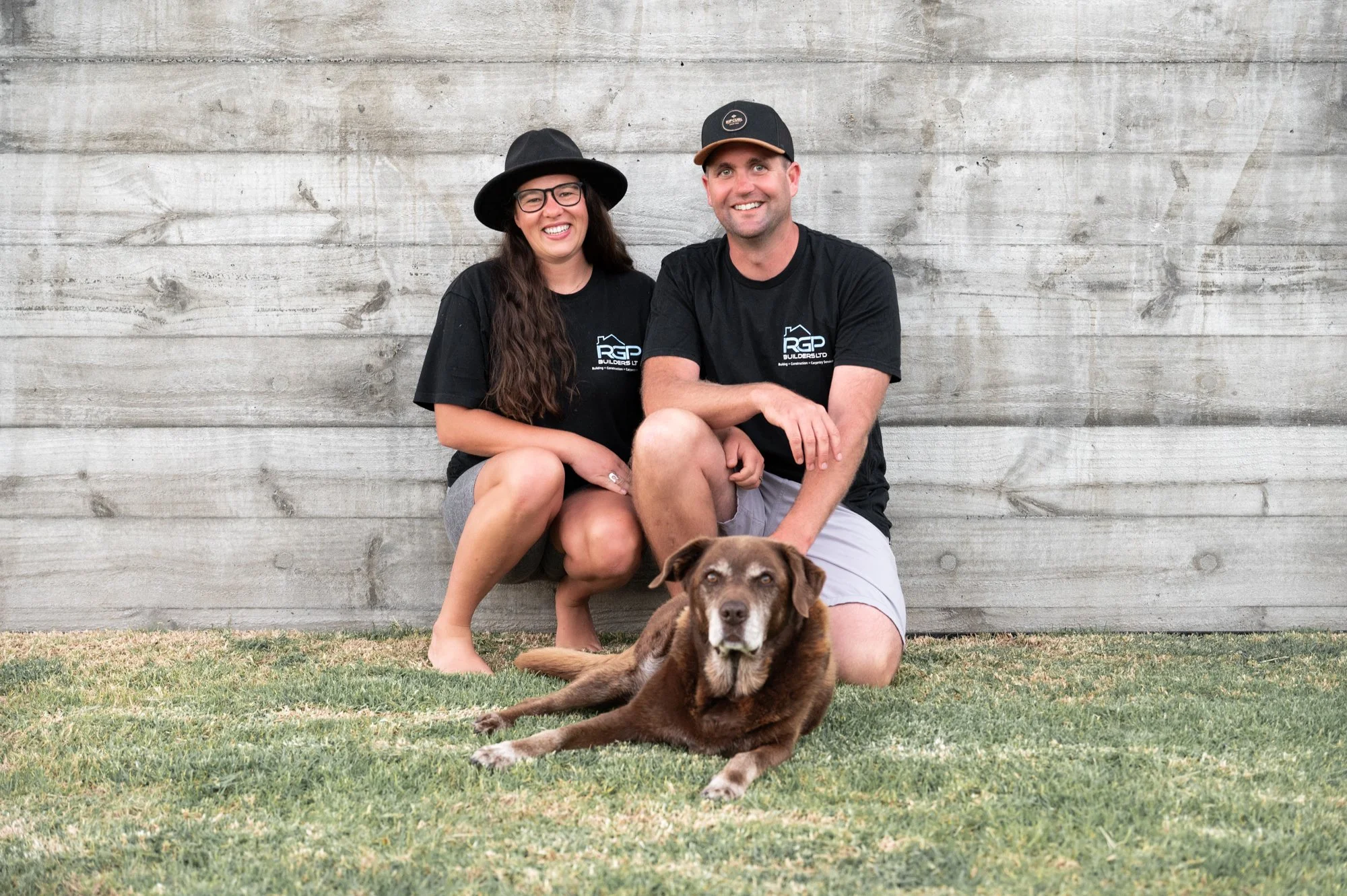 A smiling couple sitting on the grass in front of a wooden wall with their brown dog, which is lying on the grass in front of them. The woman has long dark hair, glasses, a black hat, and is wearing a black T-shirt. The man has short hair, a cap, and is wearing a black T-shirt. Both are casually dressed and appear happy.