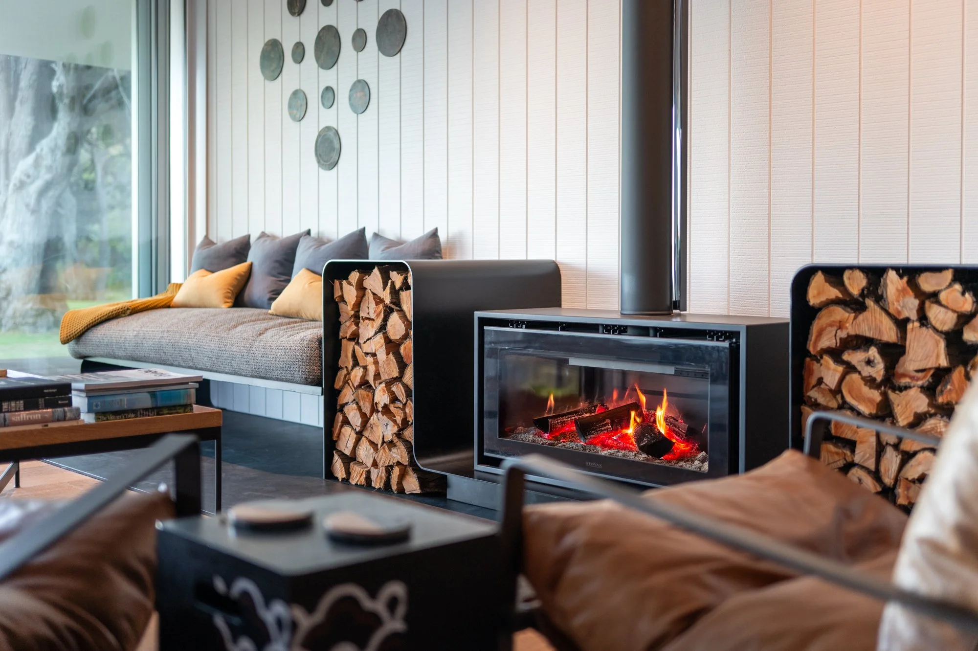 Living room with a beige couch, colorful pillows, a wood coffee table with books, and a modern fireplace with visible flames. There are two stacks of chopped firewood on either side of the fireplace, and large windows letting in natural light. A wall art piece with circular metallic plates is above the couch.