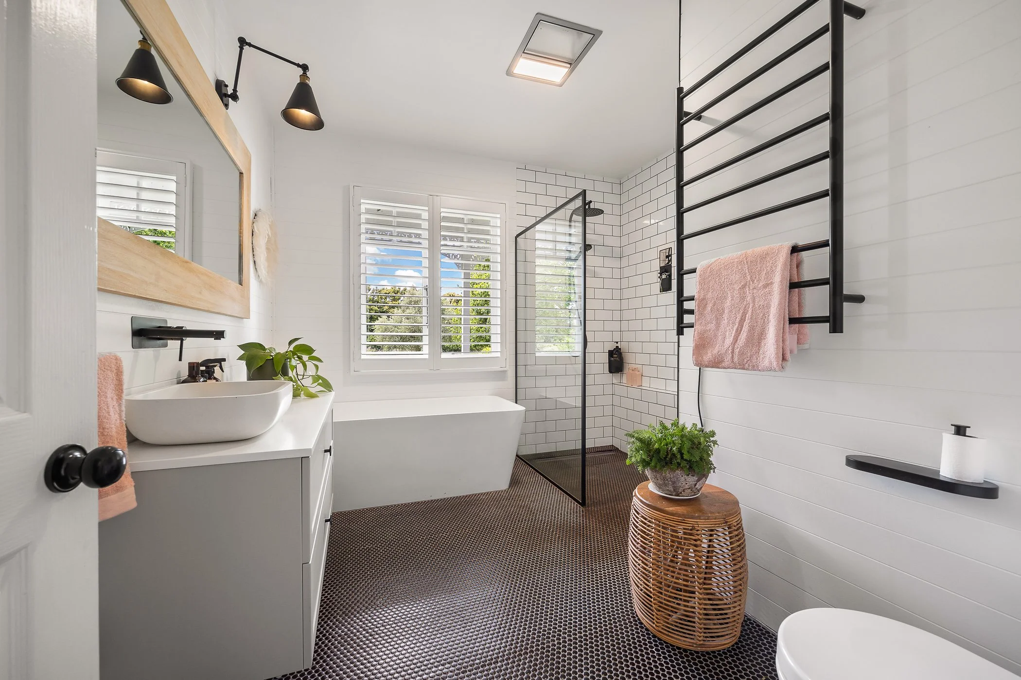 A modern bathroom with white subway tile walls, black fixtures, a walk-in shower with glass door, and a window with plantation shutters revealing a green outdoor view. There is a white vanity with a vessel sink, black faucet, pink towel, black wall-mounted lighting, a wicker stool with a plant, and a black towel rack with pink towels.