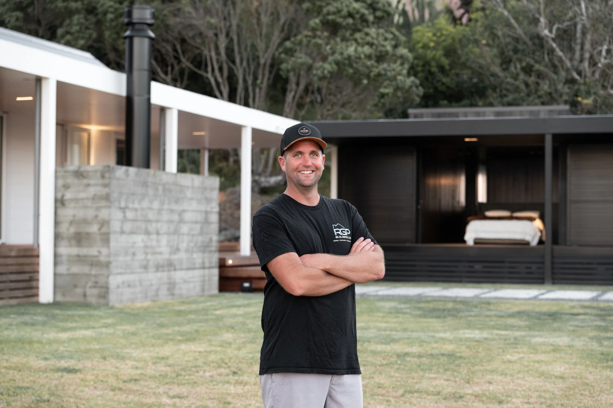 Man standing outdoors with arms crossed, smiling, wearing a black cap and black t-shirt in front of modern house with large windows, deck, and lush green trees in background.