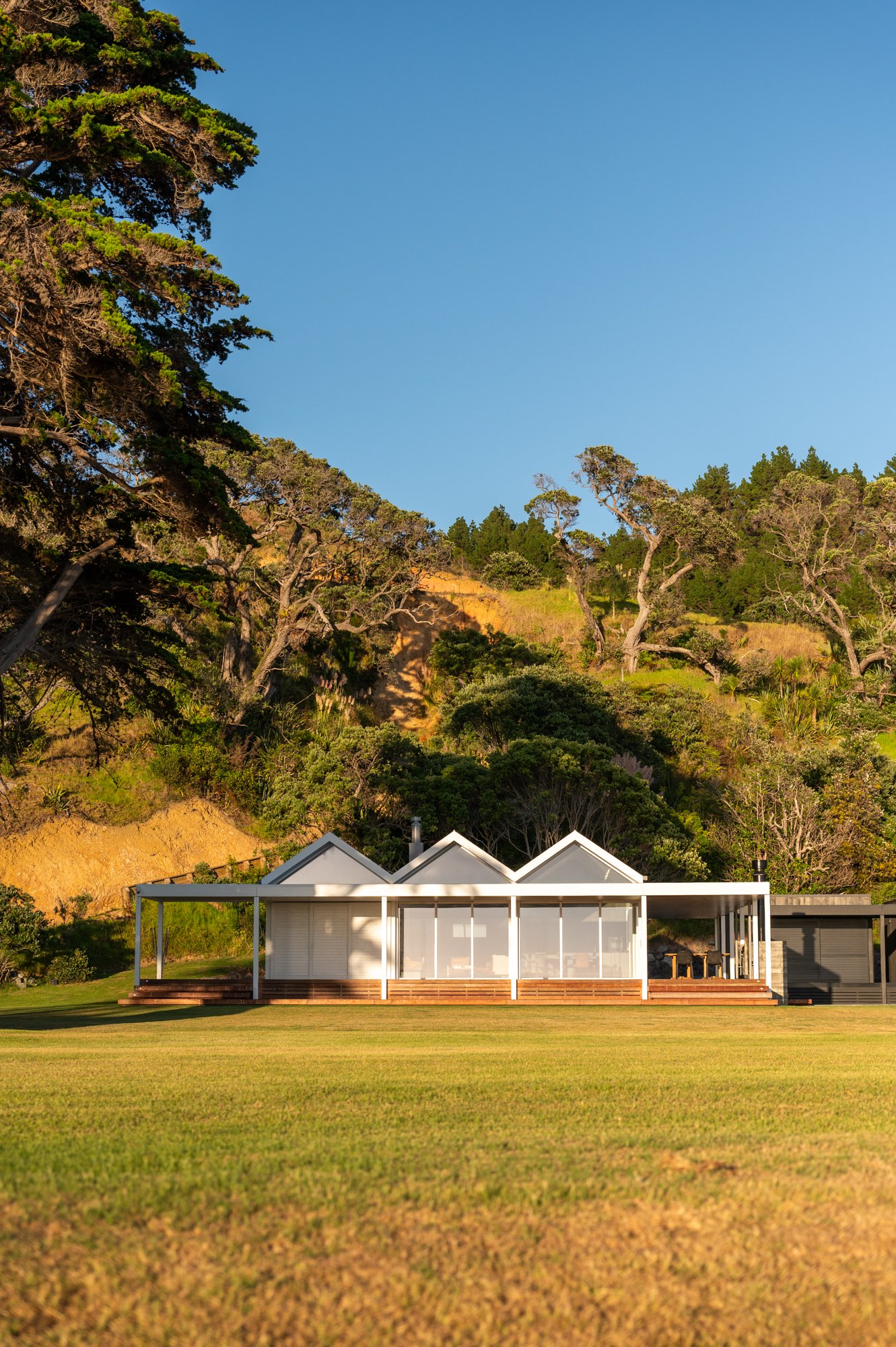 Modern white house with a screened porch, set in a green lawn with a hilly, wooded hillside behind it. The hillside has tall trees and a clear blue sky above.