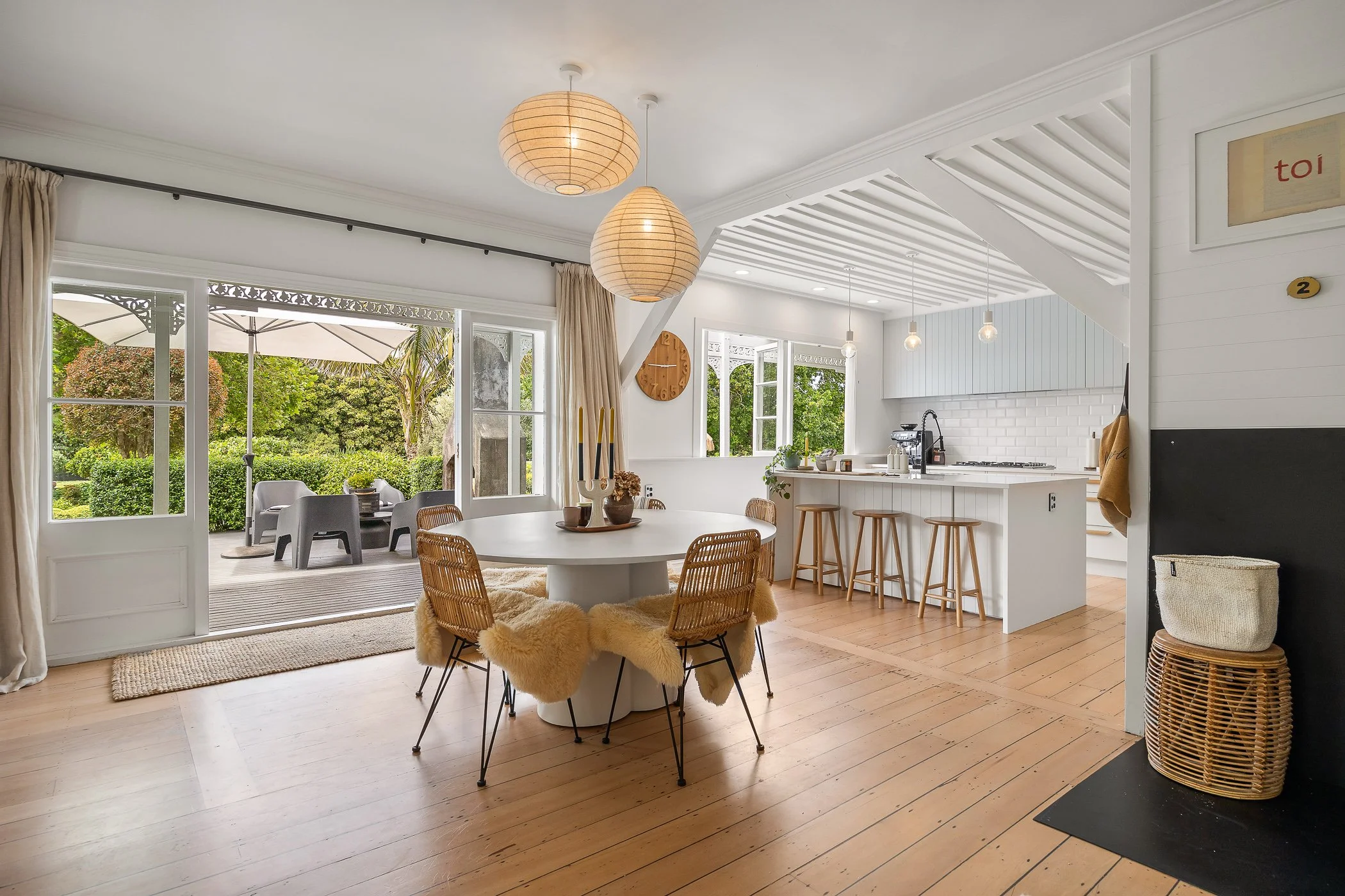Bright, modern kitchen and dining area with large sliding glass doors leading to an outdoor patio with seating and an umbrella, decorated with wooden accents, white walls, and hanging pendant lights.