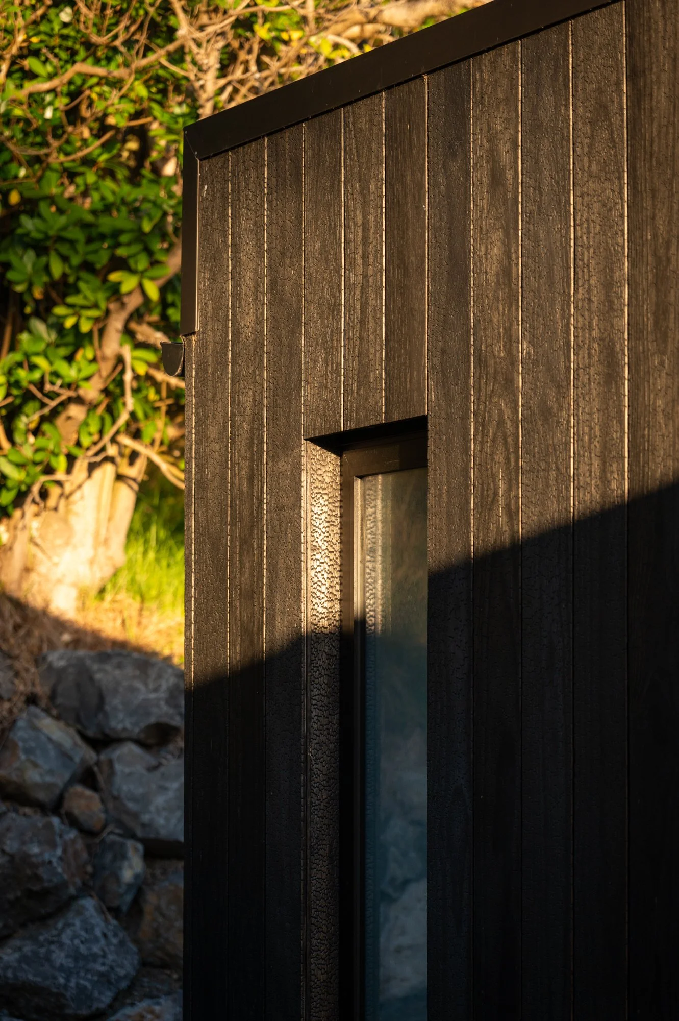 Close-up of a dark wooden exterior wall with a small window, sunlight casting shadows, and a background of green foliage and rocks.