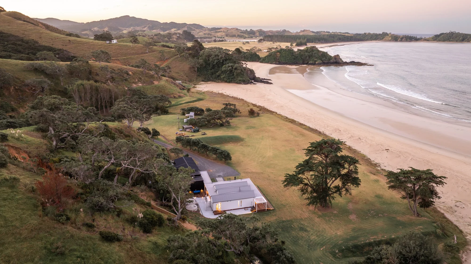 An aerial view of a coastal landscape featuring a sandy beach, rolling hills with sparse trees, a small cluster of houses including a white modern house with outdoor lighting, and the ocean with gentle waves at sunset.