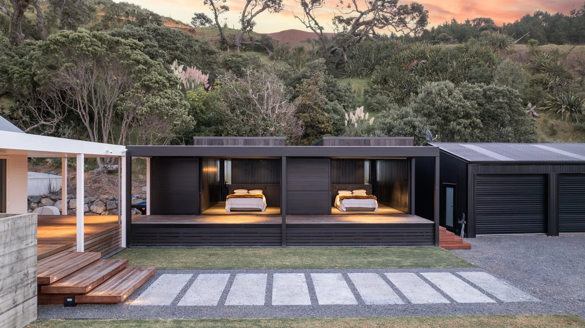 Modern black house with two outdoor bedrooms, each with a bed, overlooking a lush green hillside with trees at sunset.