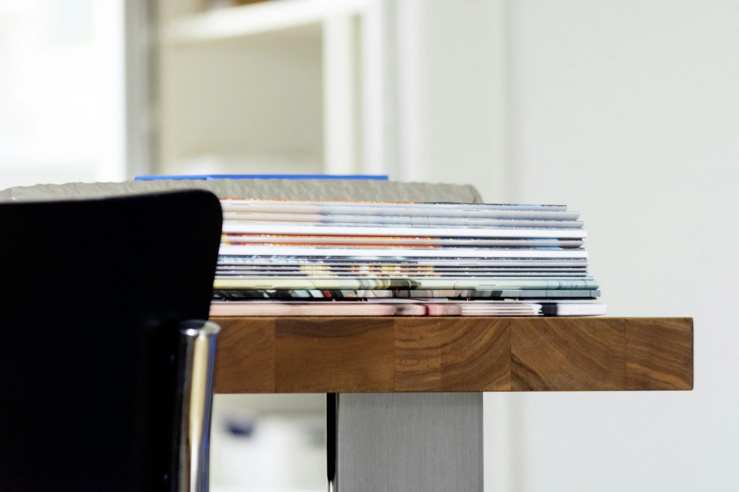 Close-up of a polished wooden table with a stack of magazines or books on it, partially obscured by a black chair back in the foreground, with a blurred light-colored wall in the background.