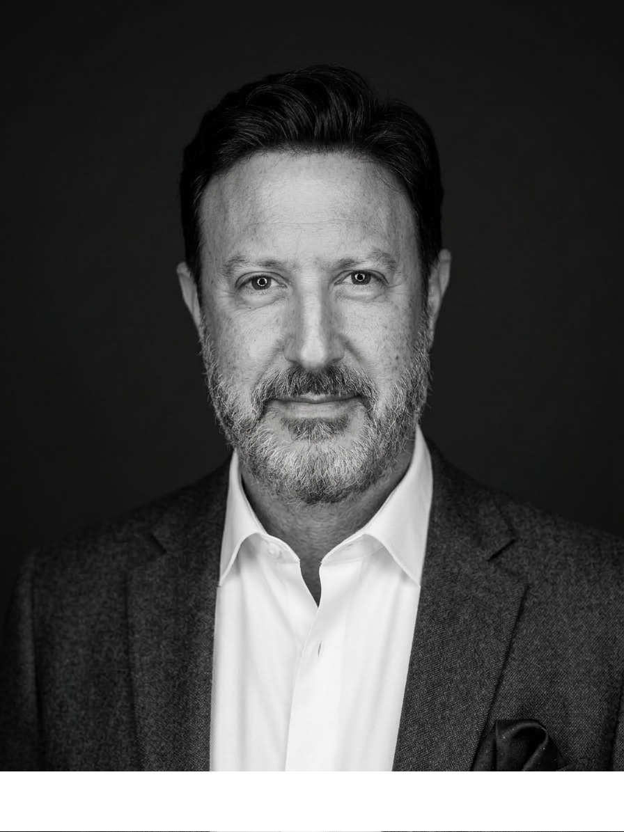 Black and white portrait of a middle-aged man with a beard, wearing a suit jacket and white shirt, against a black background.