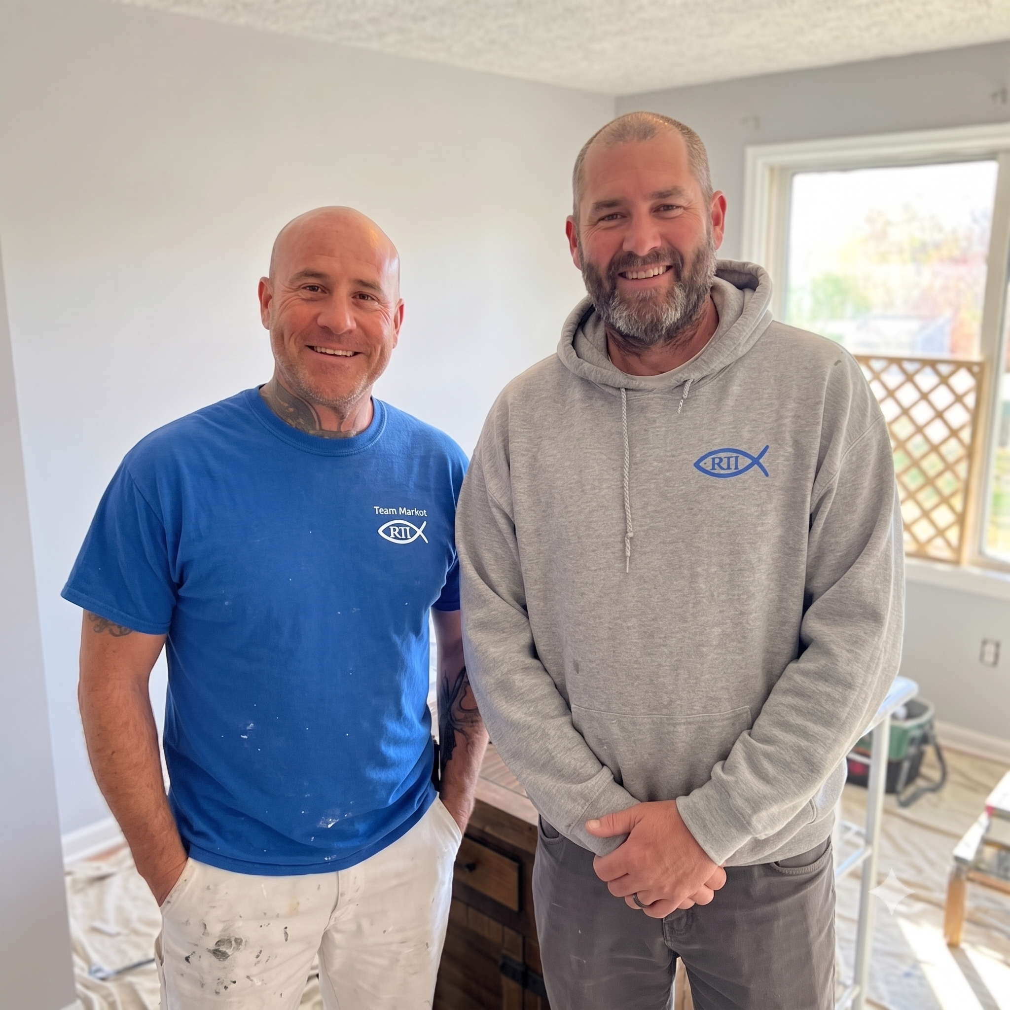 Two smiling men standing side by side indoors, one wearing a blue shirt and white pants with paint spots, the other in a gray hoodie with a logo, in a room with a window and white walls.