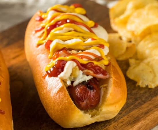 Hot dog in a bun topped with ketchup, mustard, and relish, served with potato chips on a wooden surface.