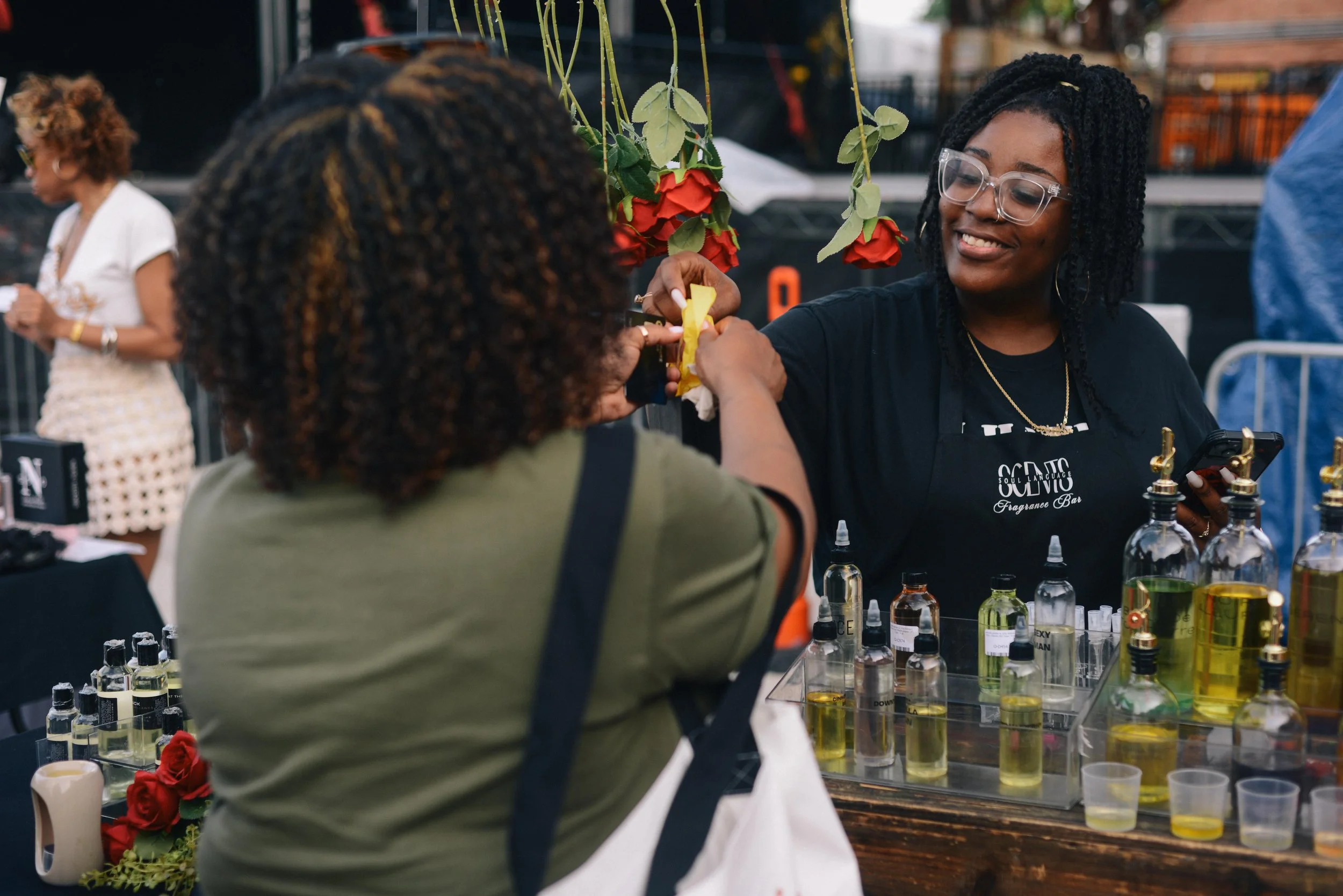 Two women at a fair or market stand, with one woman smiling and wearing glasses and a black shirt, handing a yellow item to the other woman, who has curly hair and is wearing a green jacket. The stand displays various bottles of colorful liquids, and there are hanging red roses and flowers above the stand.