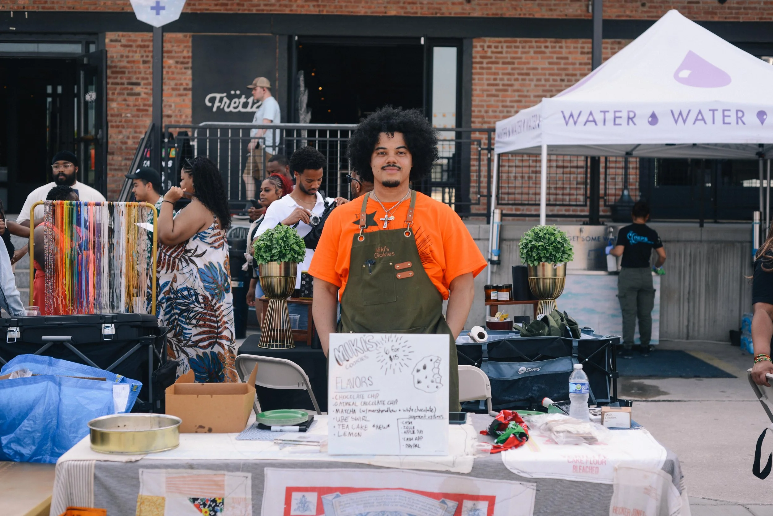 A young man with curly hair, wearing an orange shirt and a green apron, standing behind a table at an outdoor market or fair. The table has various items including potted plants and a sign advertising cookies. Several people are visible in the background, some browsing and others working at different booths. A white tent with purple lettering and a purple drop symbol is also visible.