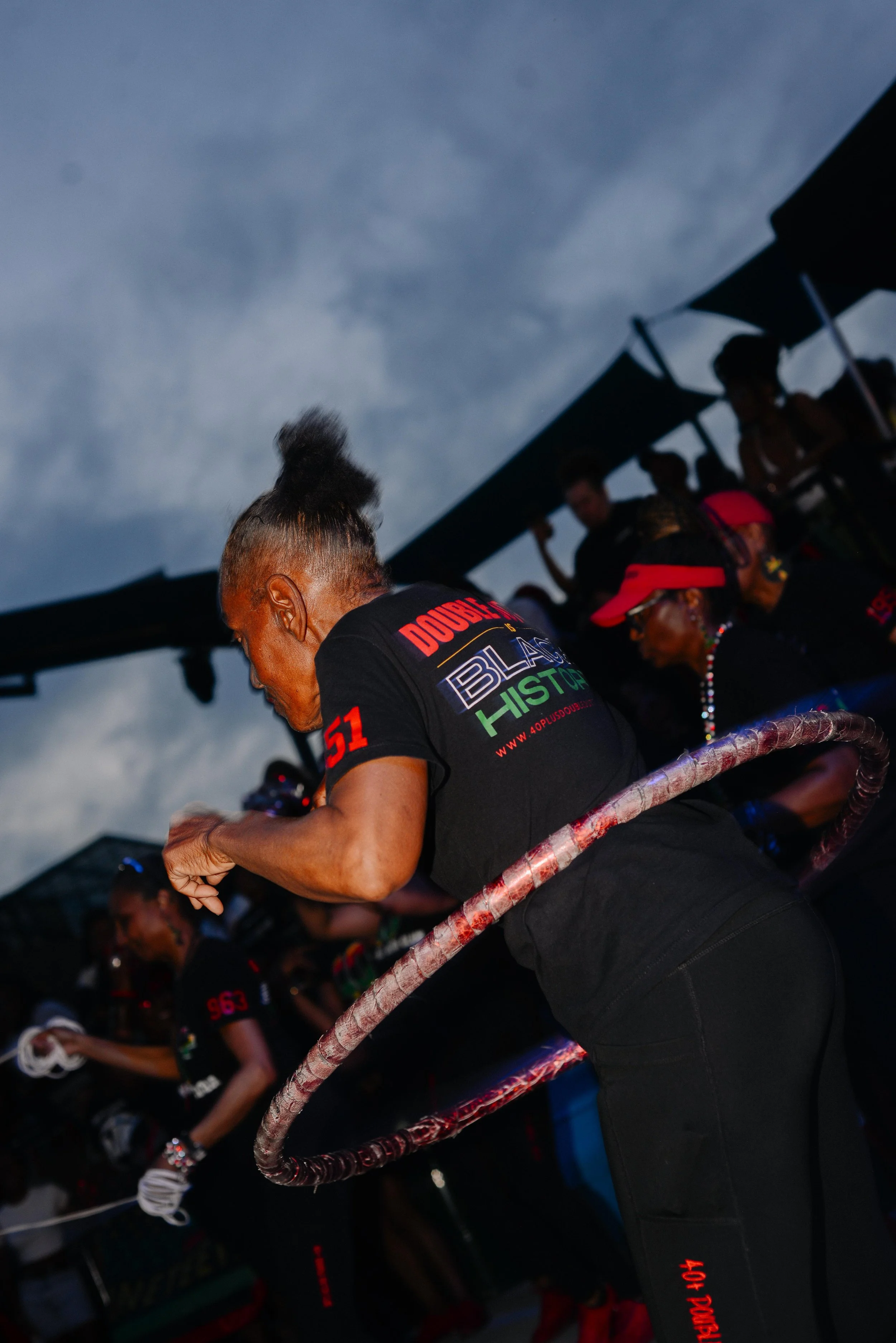 A group of people gathered under a canopy at dusk, some wearing racing gear, with a woman in the foreground adjusting her gloves.