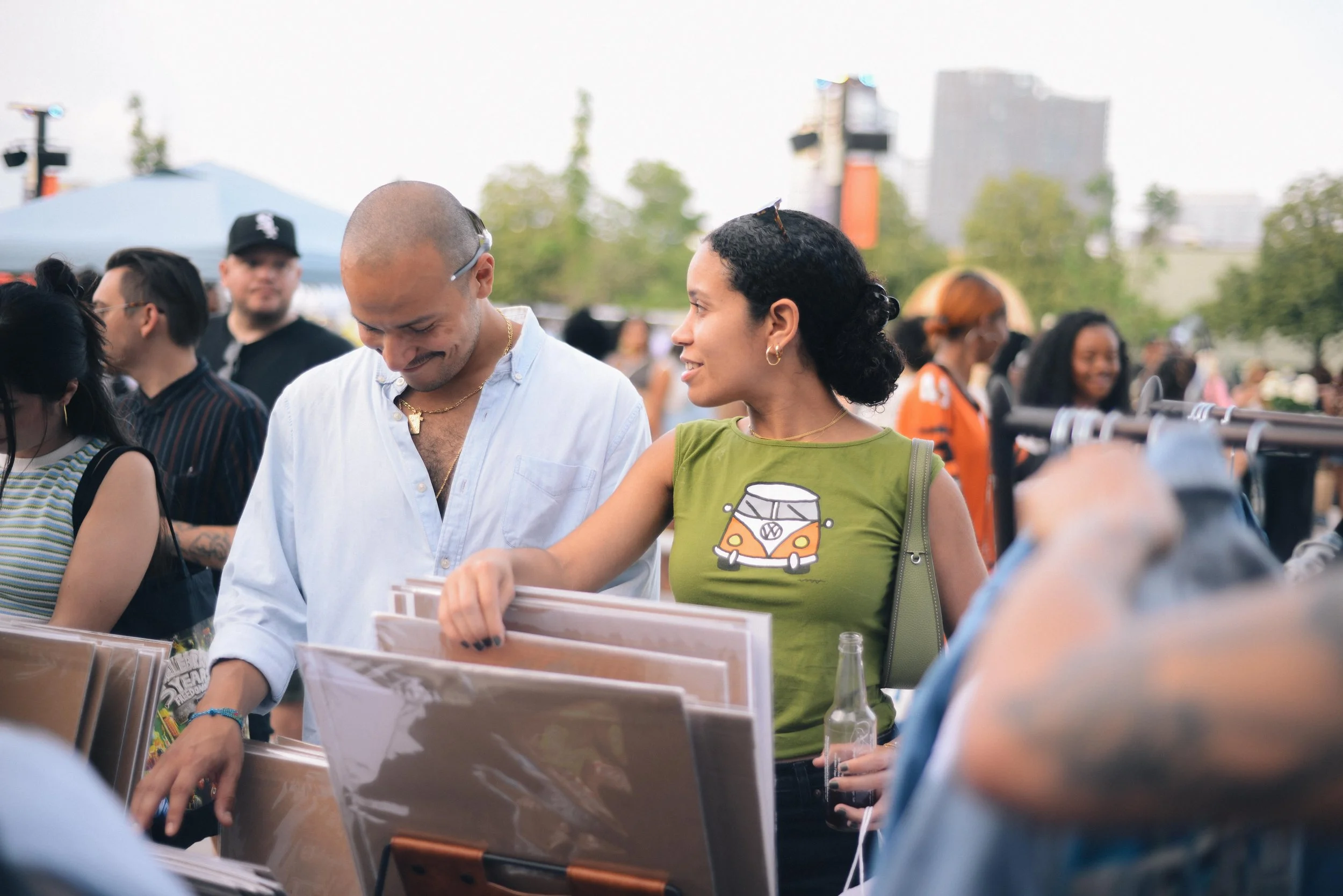 A diverse group of people at an outdoor event, browsing through vinyl records at a market stall. The background shows trees, a tent, and a city skyline.