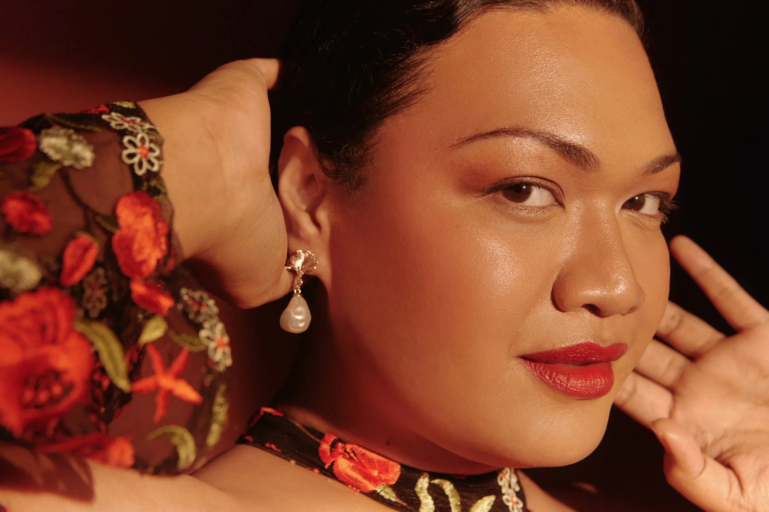Close-up of a woman with smooth skin, dark hair, red lips, wearing a pearl earring and an embroidered garment, with one hand near her face against a dark background.