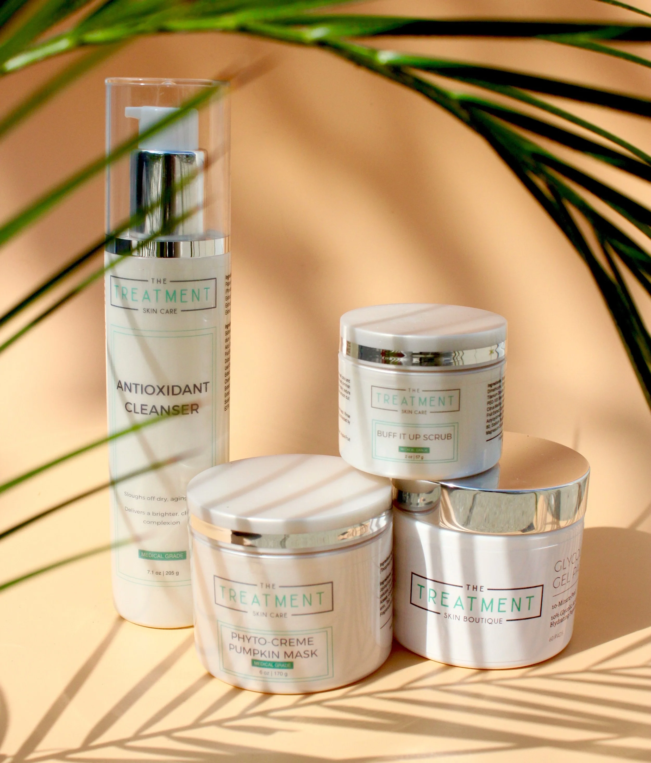 Collection of skincare products on a white surface with palm leaf shadows in the foreground, including a tall bottle labeled Antioxidant Cleanser, and smaller jars labeled Pumpkin Mask, Buff It Up Scrub, and Glycolic Gel.