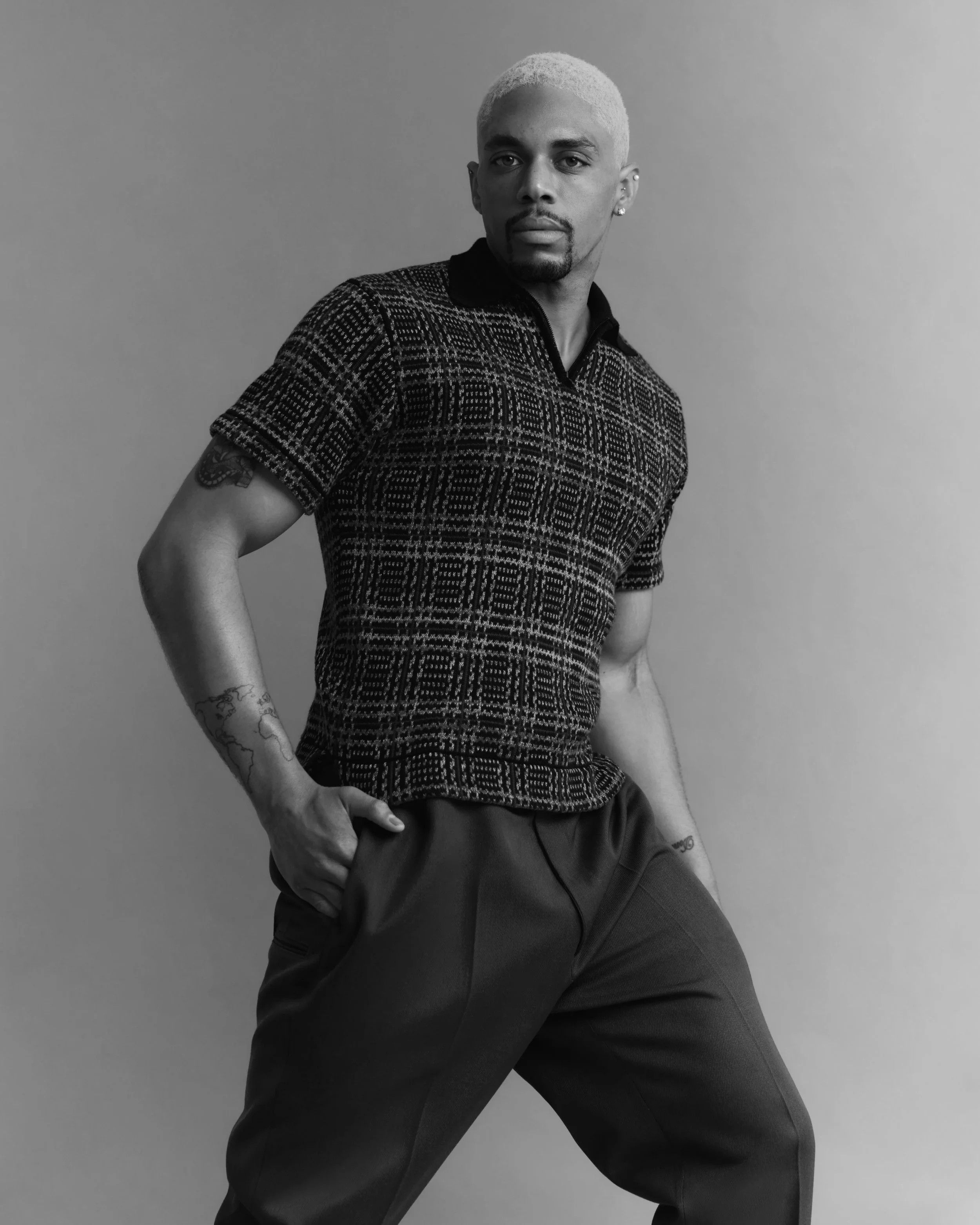 Black and white portrait of a person with short, light-colored hair, wearing a patterned shirt and dark pants, striking a confident pose against a plain background.