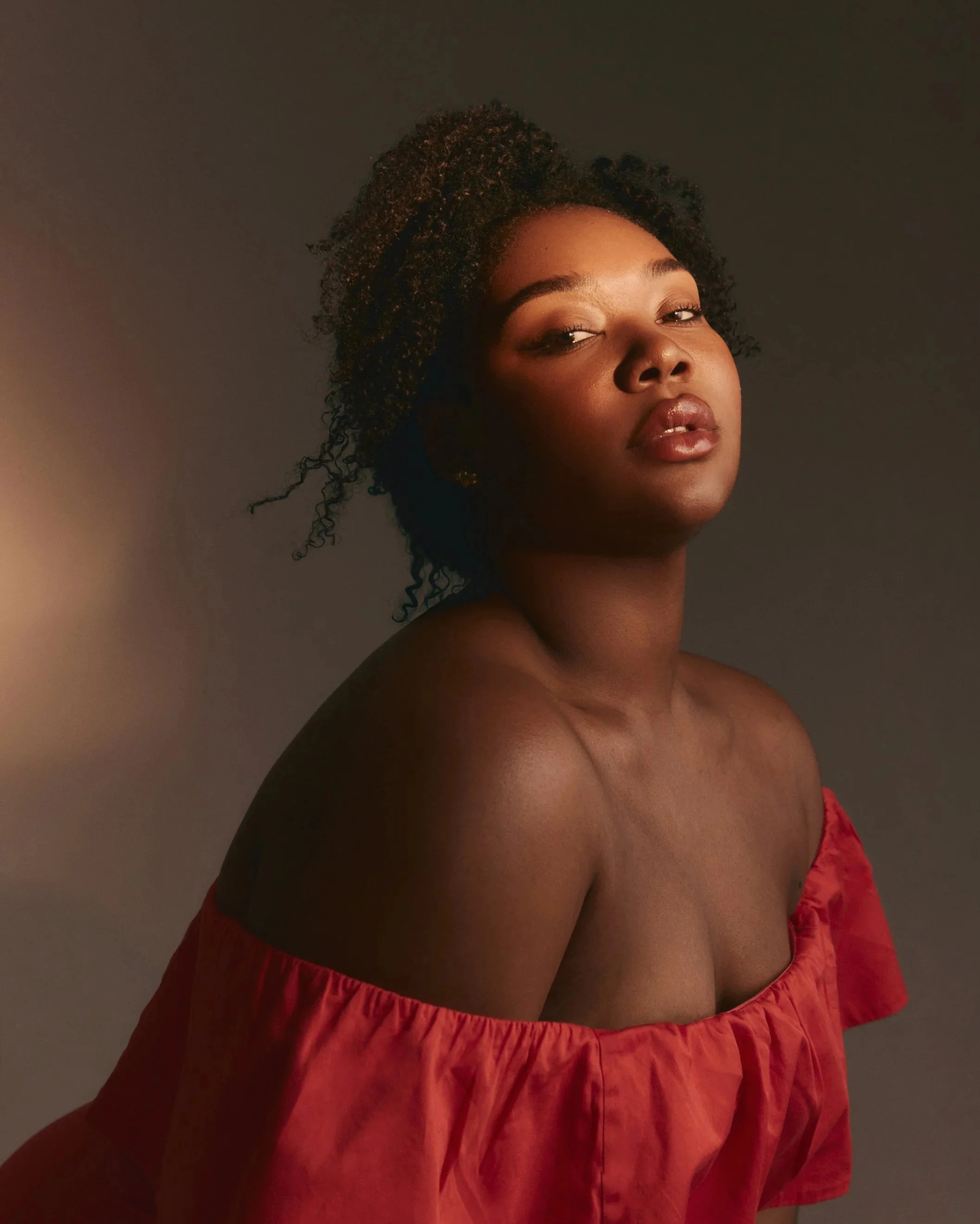 A woman with dark skin and curly hair wearing a red off-the-shoulder top, looking slightly to the side with a neutral expression against a dark background.