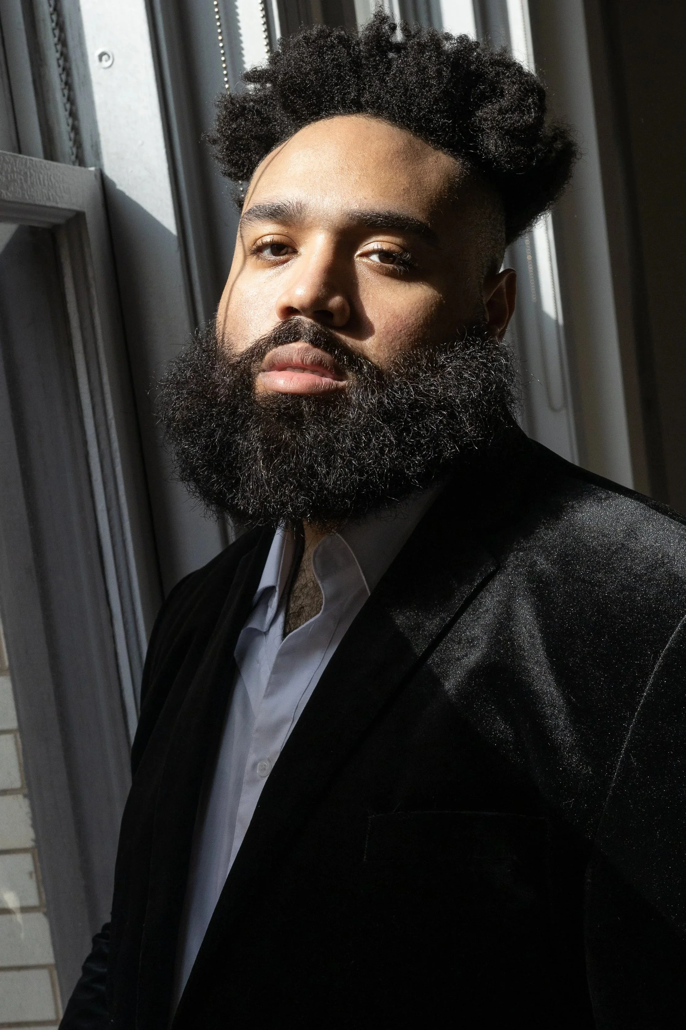 A man with dark skin, curly hair, and a beard wearing a black blazer over a white shirt, is standing near a window with sunlight illuminating his face.