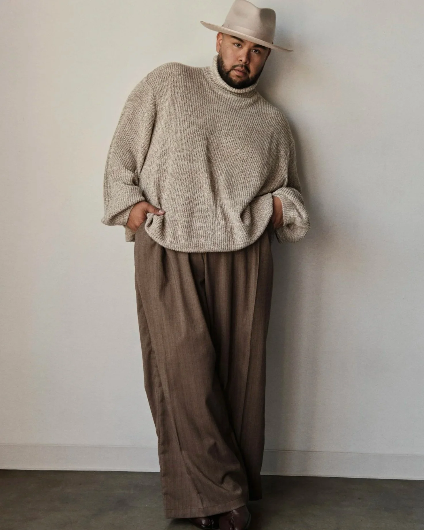 A man stands against a plain wall, wearing a large beige fedora, a beige knitted turtleneck sweater, brown wide-leg trousers, and dark brown shoes. His hands are in his pockets, and he gazes directly at the camera.
