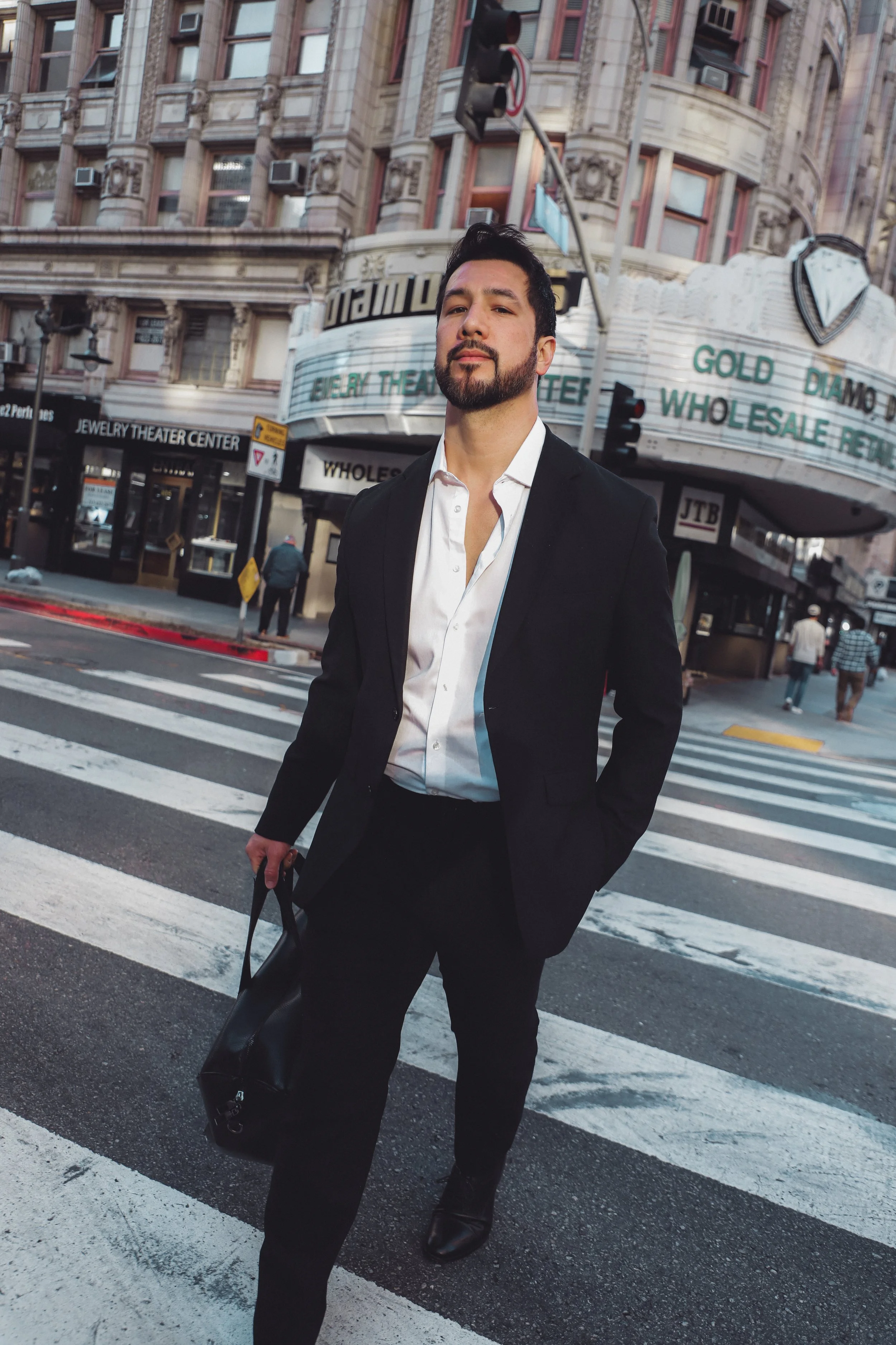 A man dressed in a black suit and white shirt walking across a crosswalk in an urban area, holding a black bag. Tall buildings and storefronts are visible in the background, including a jewelry theater center and a marquee with electronic text.