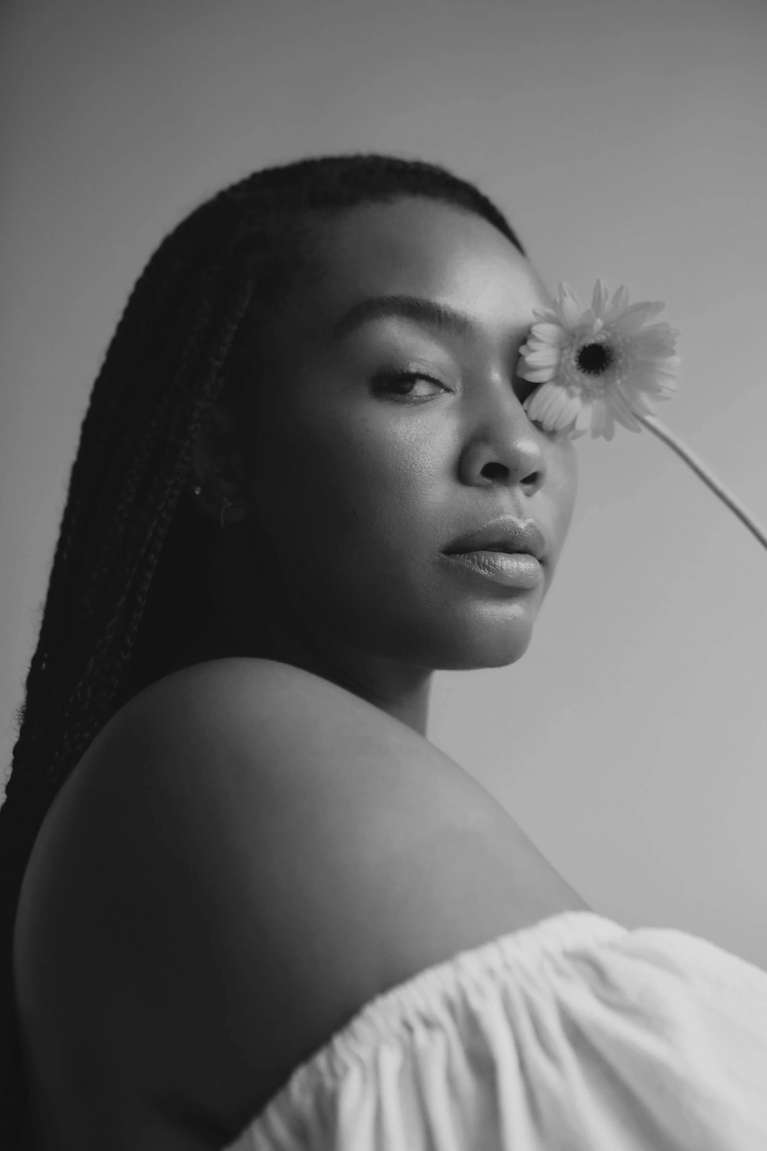 A black and white photograph of a woman with short hair holding a daisy in front of her right eye, while looking slightly to the side. She has smooth skin and well-defined eyebrows, wearing a textured, off-the-shoulder top.