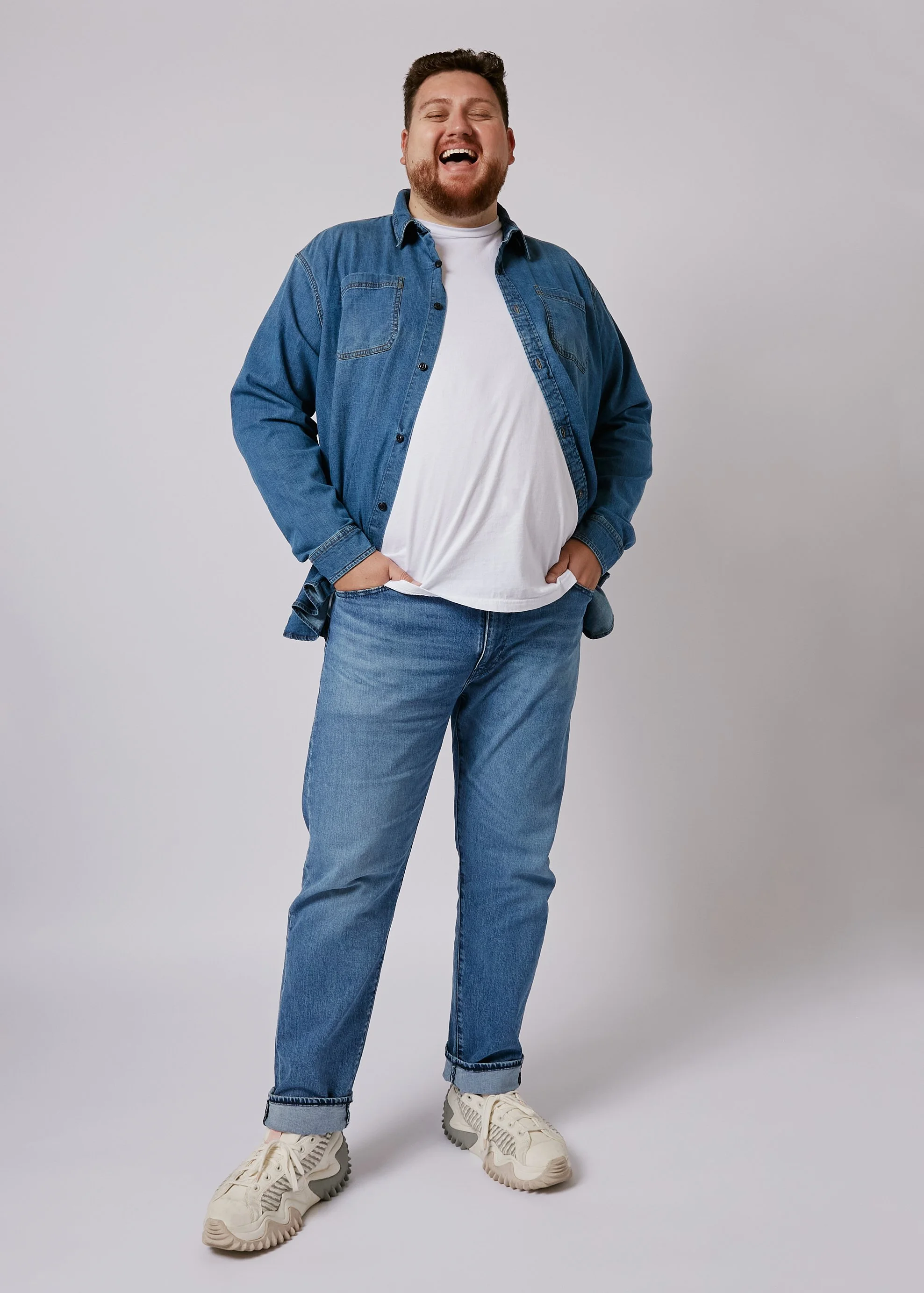 A man smiling and standing with hands in pockets against a plain gray background, wearing a denim jacket, white t-shirt, jeans, and sneakers.