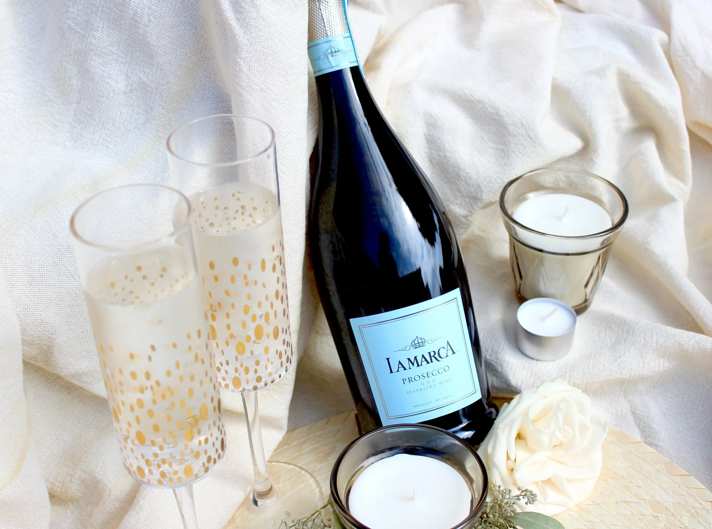 A bottle of Lamarca Prosecco with a white label and blue accents, surrounded by two empty champagne flutes with gold polka dots, a candle in a glass holder, white roses, and a white cloth background.