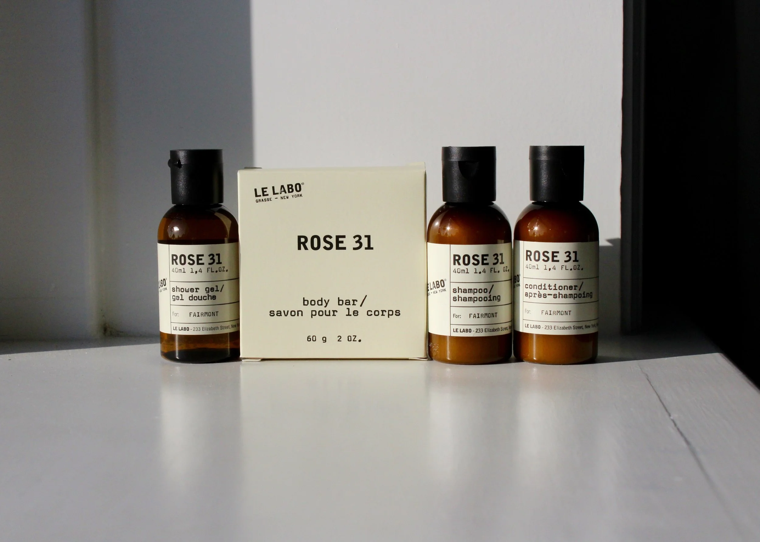 Three brown bottles labeled 'Rose 31' and a white box with the same label, placed on a white shelf against a plain wall. The bottles are for shower gel, shampoo, and conditioner.