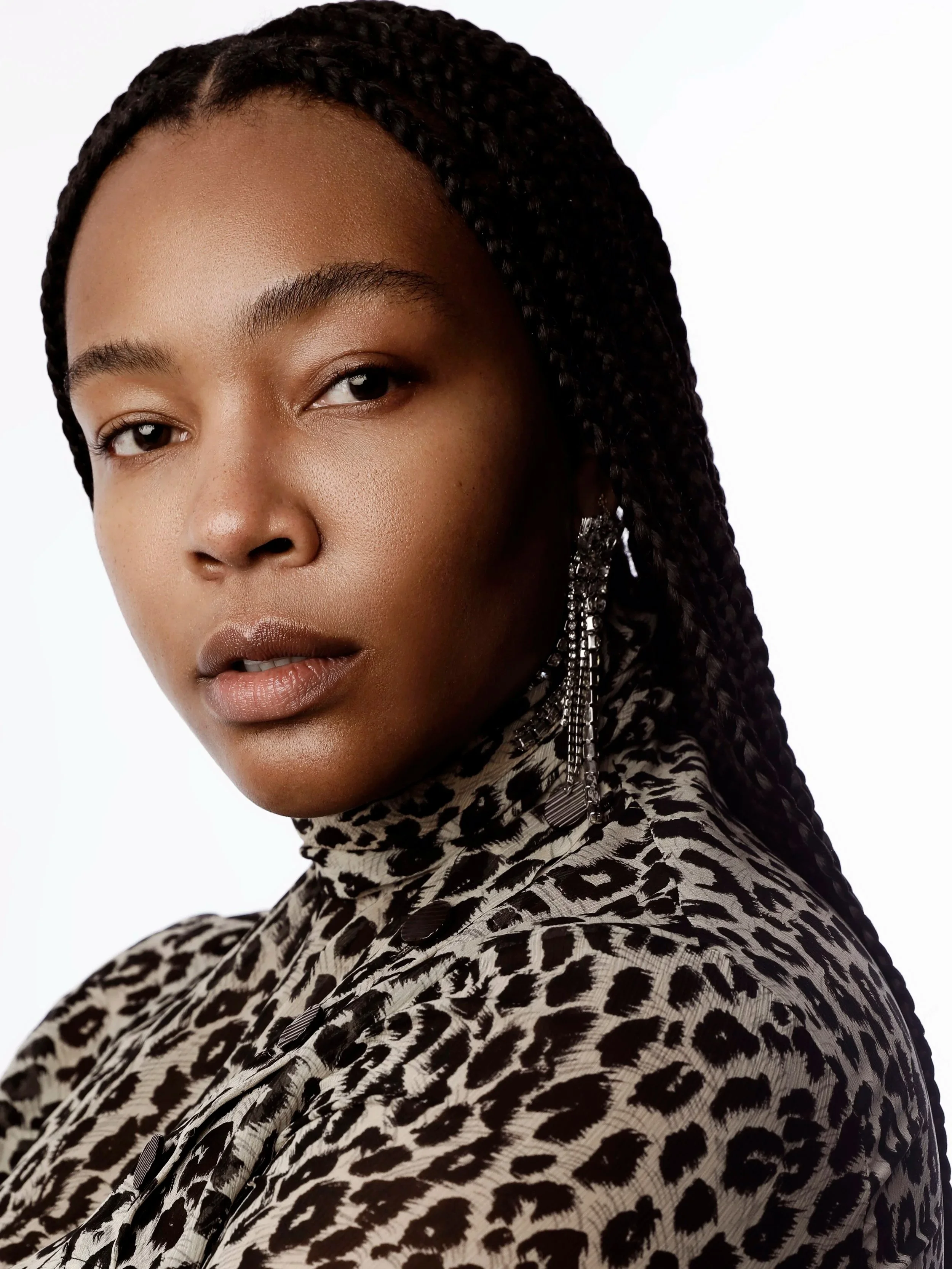 Close-up of a woman with dark skin and long braided hair wearing a leopard print scarf and earrings against a white background.