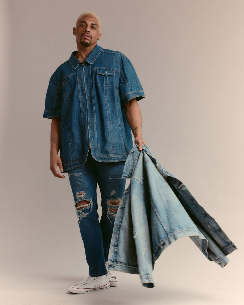 A person with blonde hair and a goatee standing against a plain background, wearing a denim shirt, ripped jeans, and holding multiple denim jackets.
