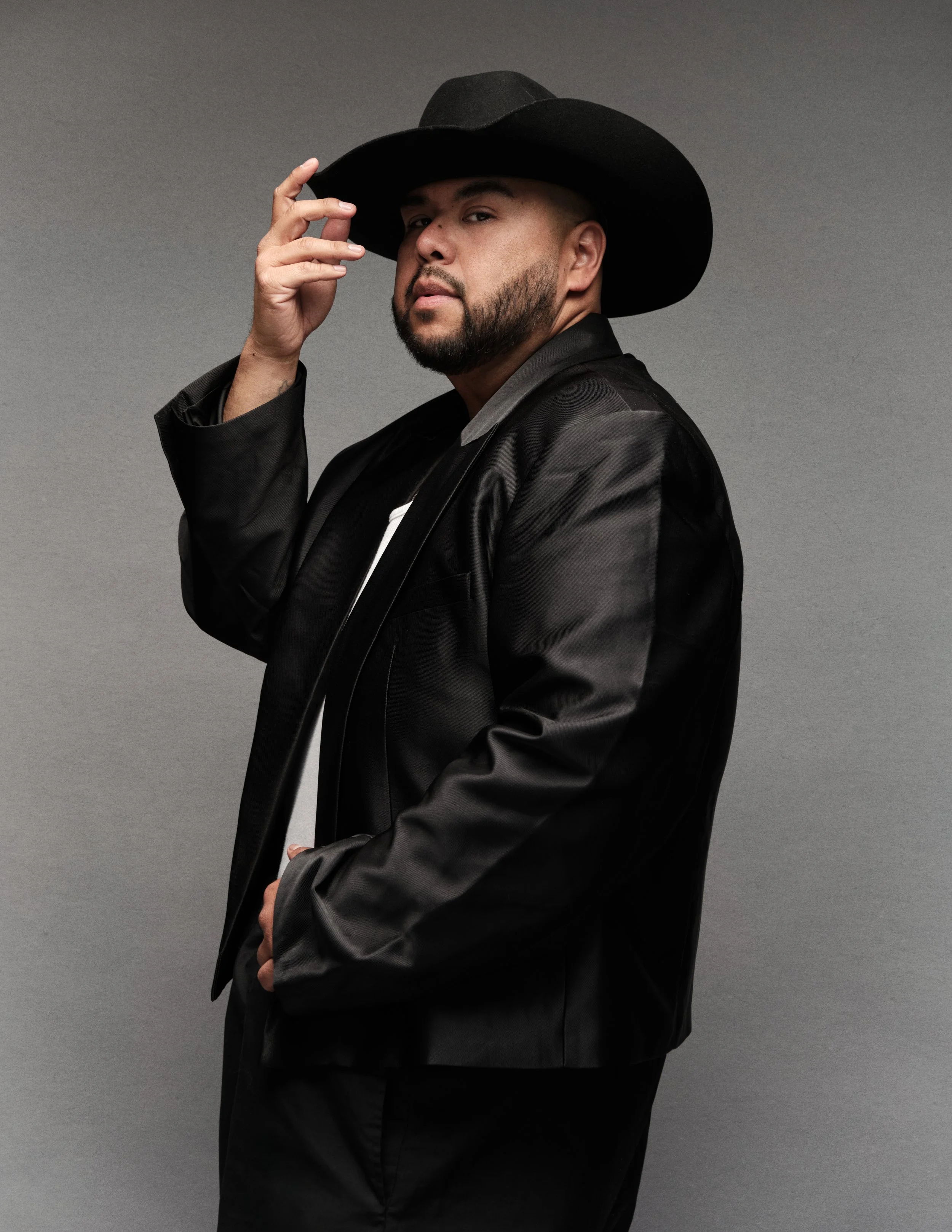 A man wearing a black cowboy hat, black satin jacket, and black shirt, posing with one hand touching the brim of his hat, against a neutral gray background.