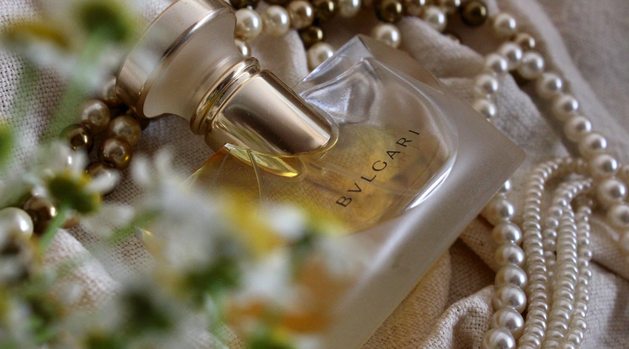 Close-up of a Bvlgari perfume bottle surrounded by pearl necklaces and a fabric background, with blurred flowers in the foreground.