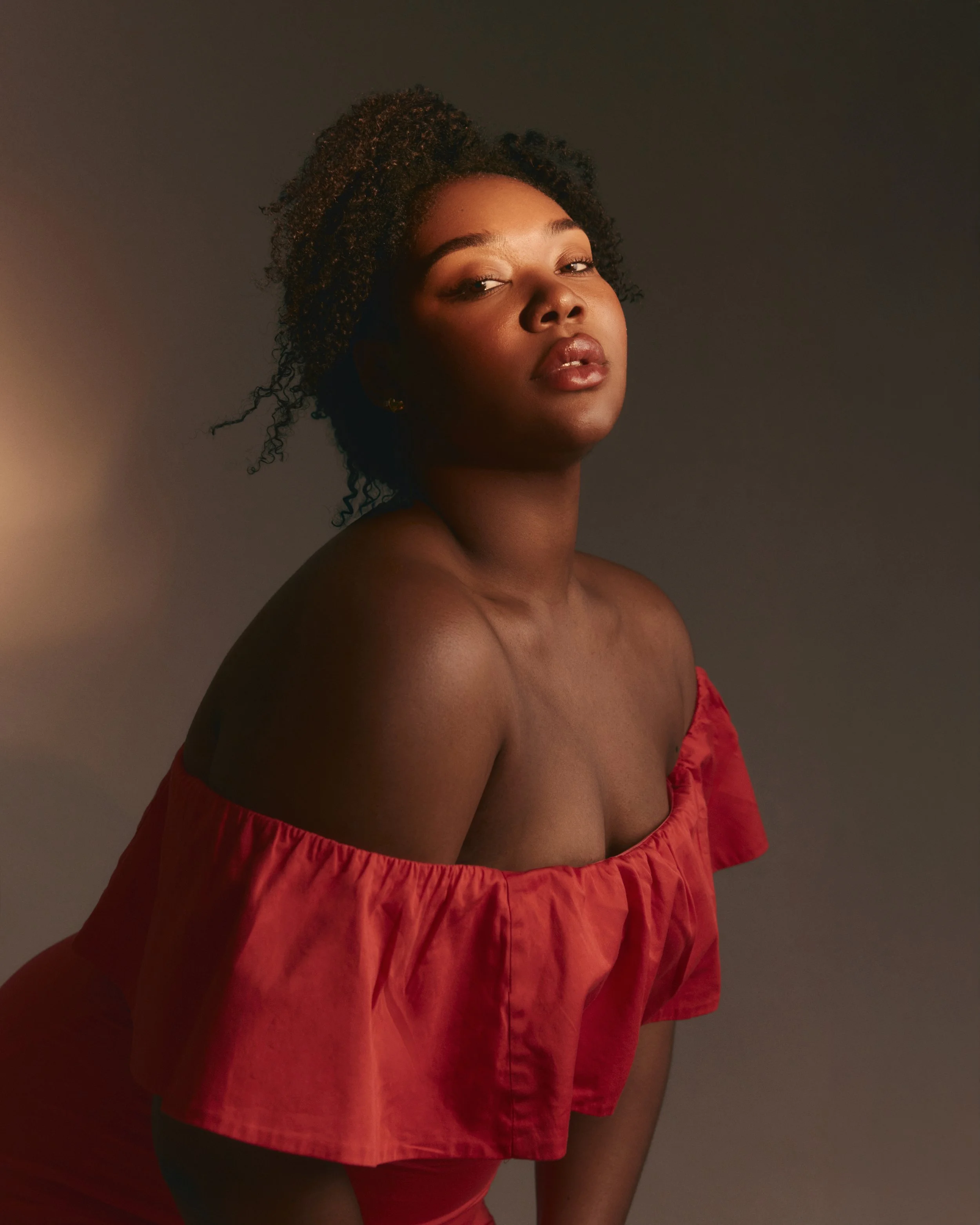 A woman with dark curly hair and brown skin wearing a red off-shoulder dress with ruffled edges, looking towards the camera against a neutral background.