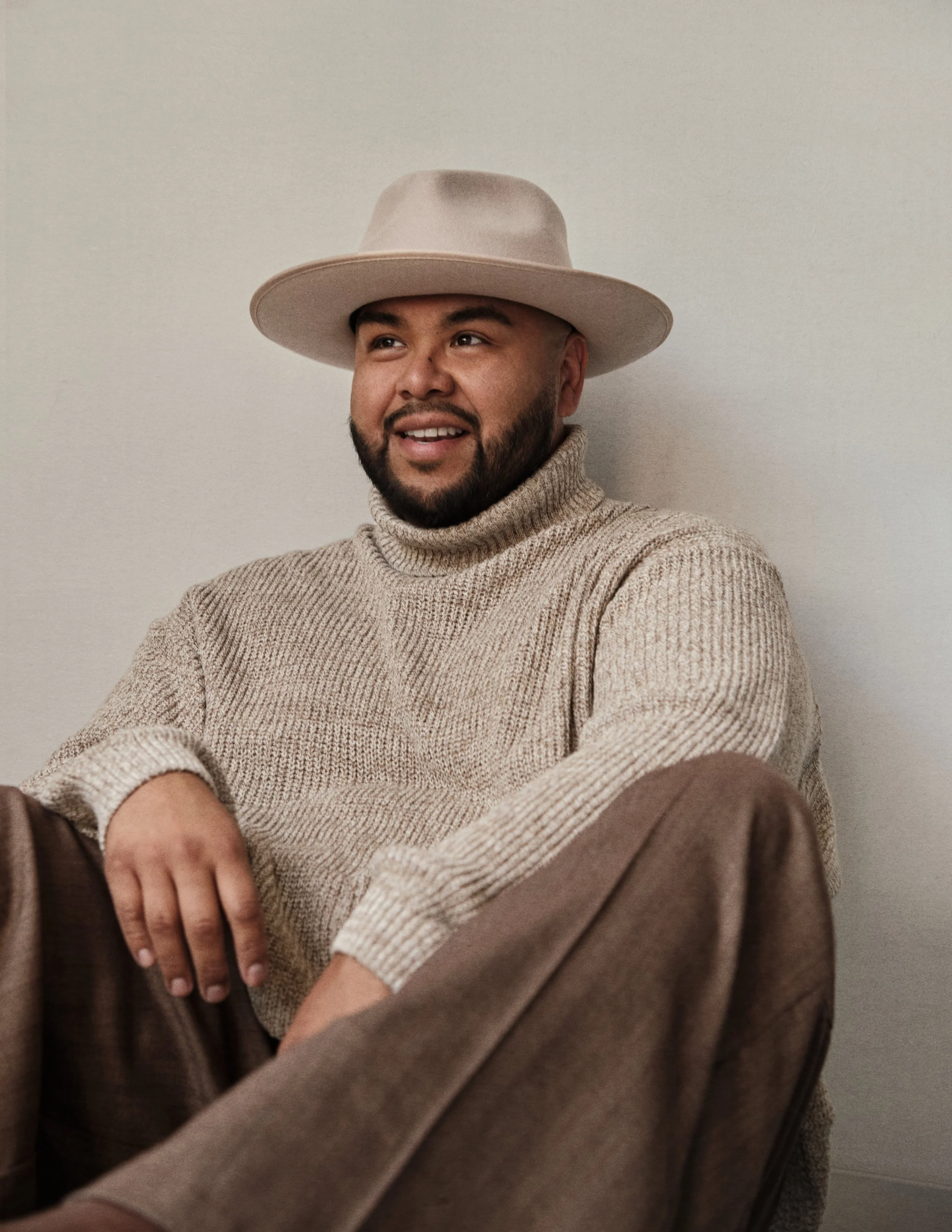 A man with a beard wearing a beige wide-brimmed hat, a beige turtleneck sweater, and brown pants sitting against a plain white wall, smiling and looking slightly to the side.
