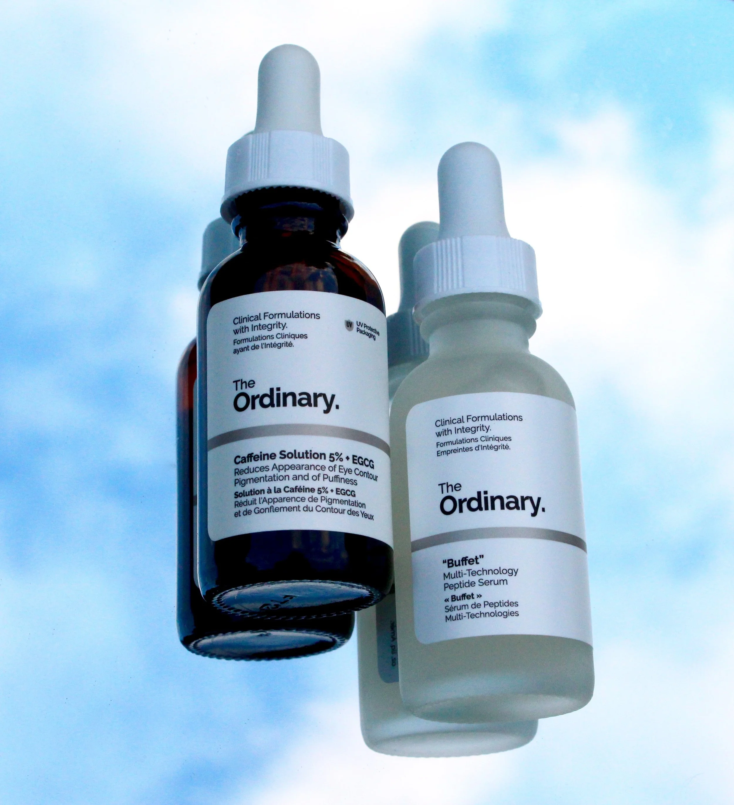 Pharmacological bottles of skincare serum with white caps, one with a label reading 'The Ordinary' and the other with text in French, set against a light blue background.