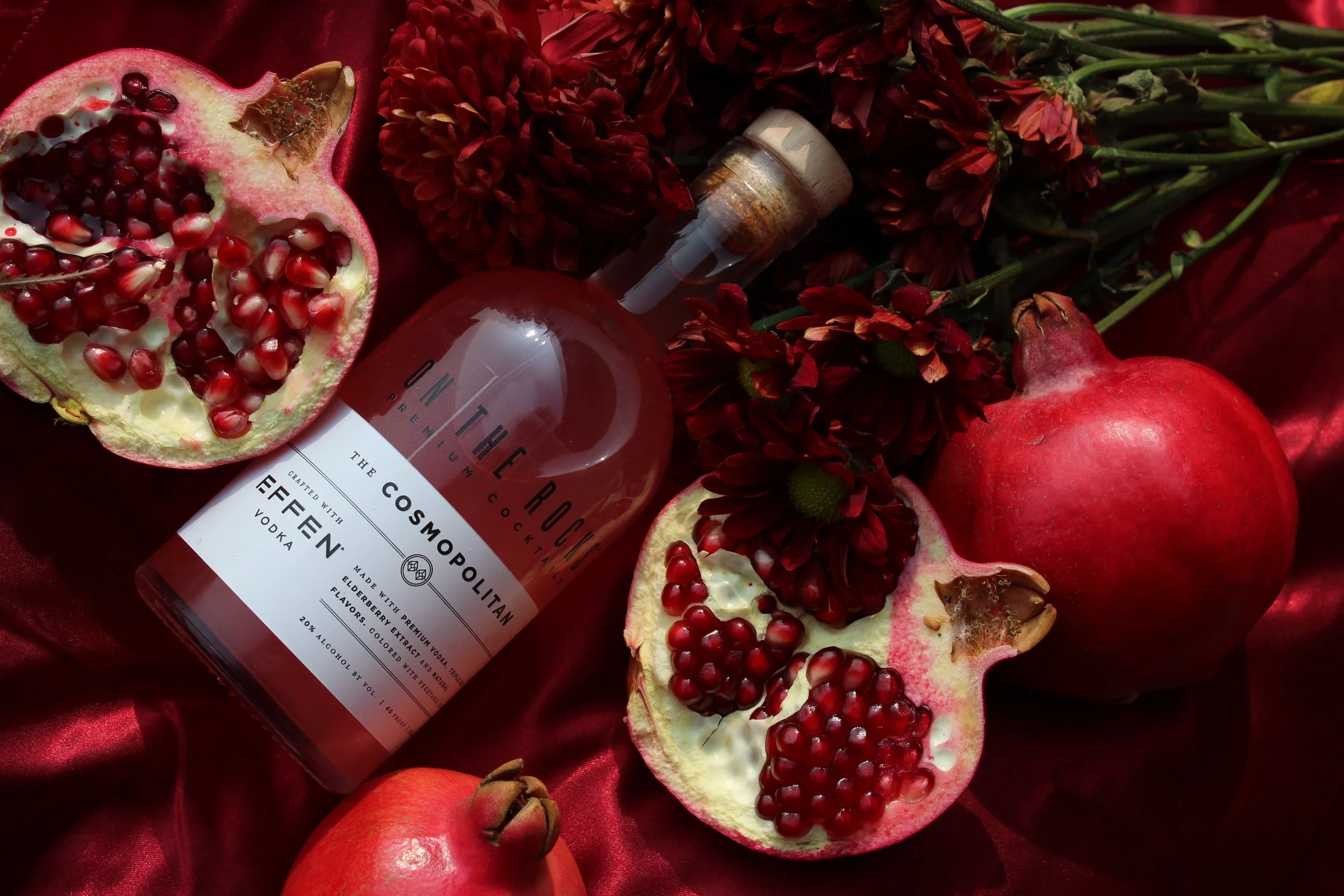 A bottle of vodka labeled 'The Cosmo' surrounded by pomegranate halves, whole pomegranates, and red flowers on red satin fabric.