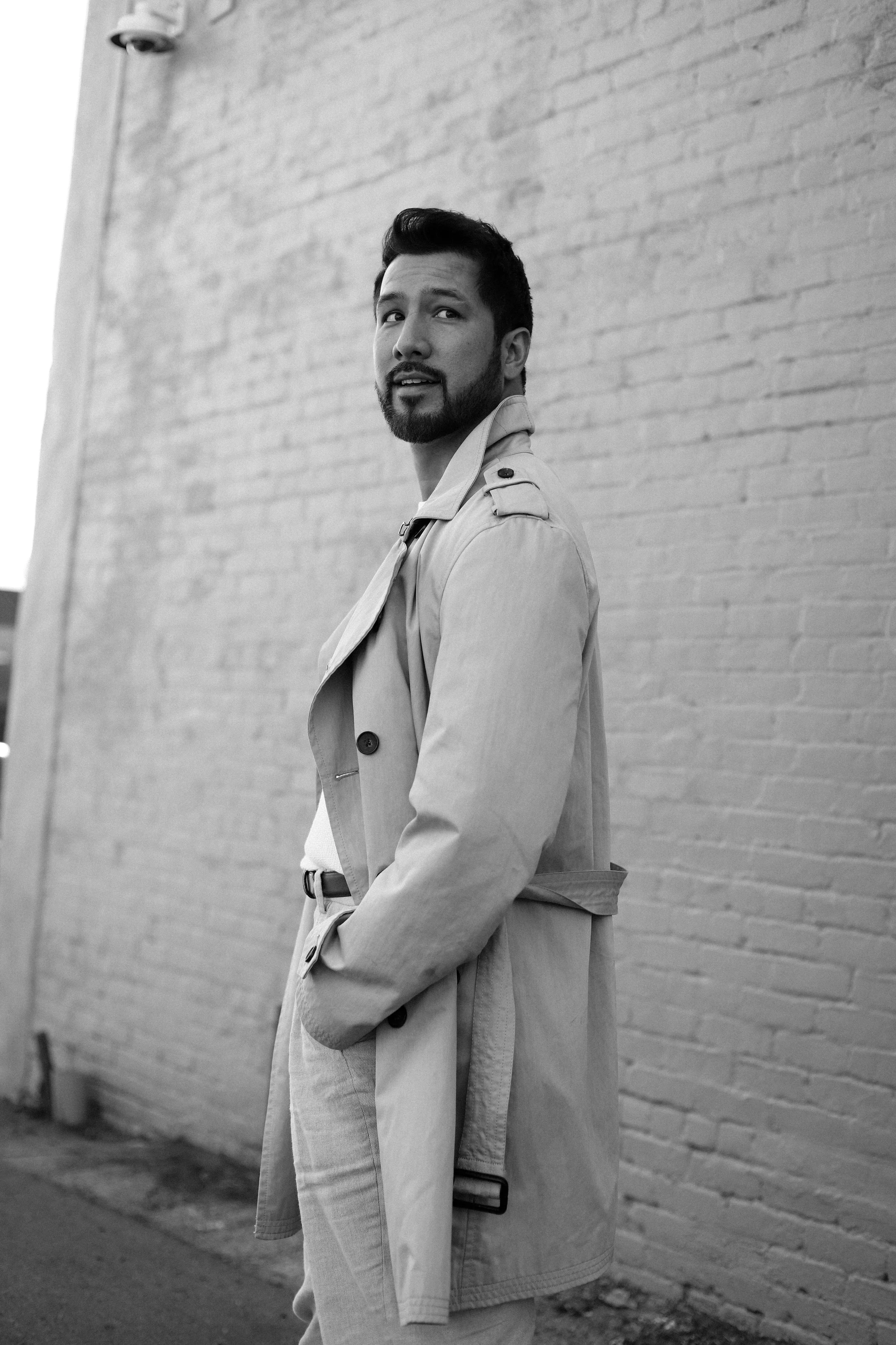 A man with a beard and mustache wearing a trench coat and light-colored pants, standing against a brick wall and looking over his shoulder.