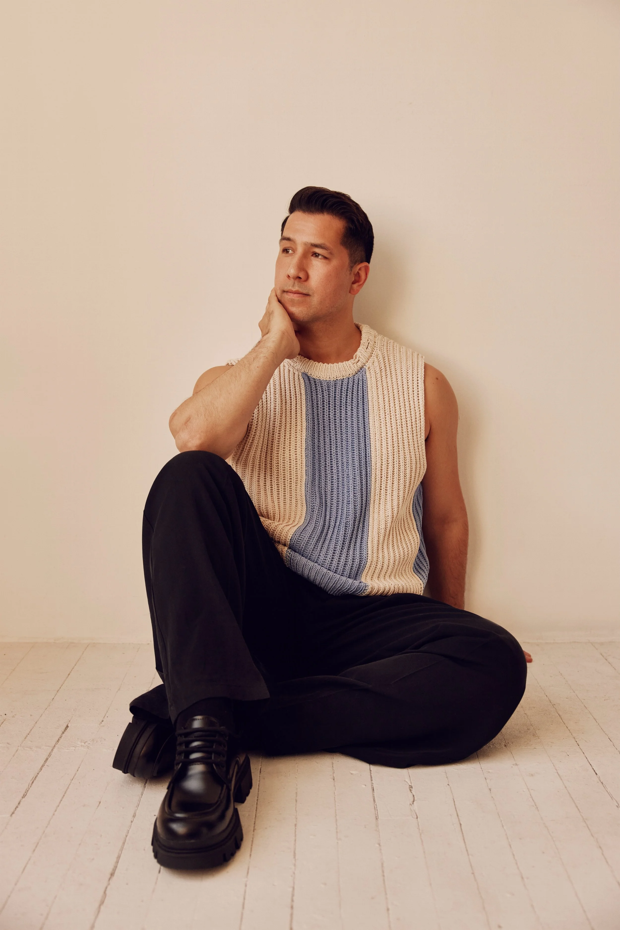 A young man sits on the floor against a plain white wall, wearing a sleeveless knit sweater, black pants, and chunky black boots. He rests his chin on his hand, gazing thoughtfully.