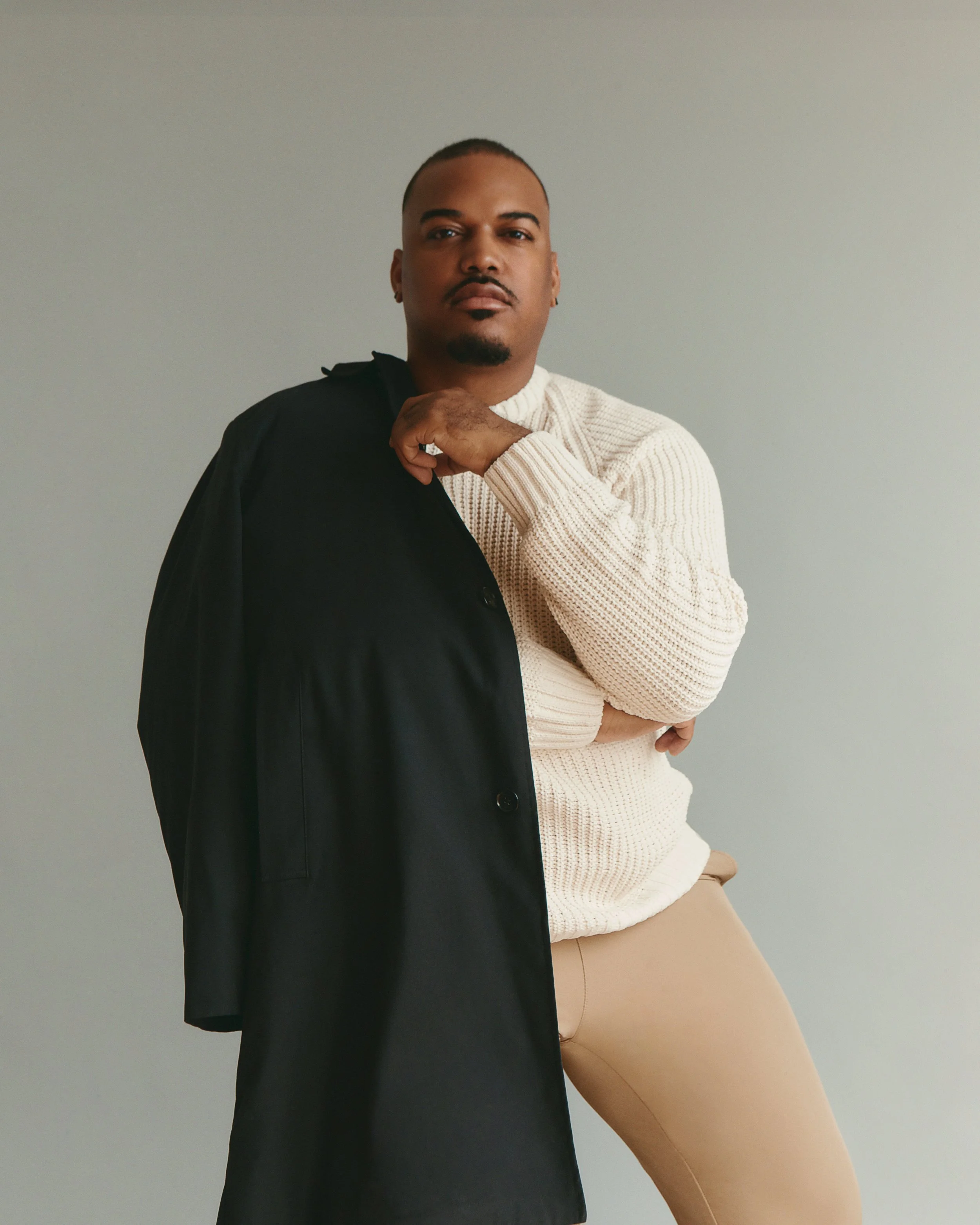 A person with a short haircut and a goatee, wearing a beige sweater and beige pants, standing against a plain gray background.