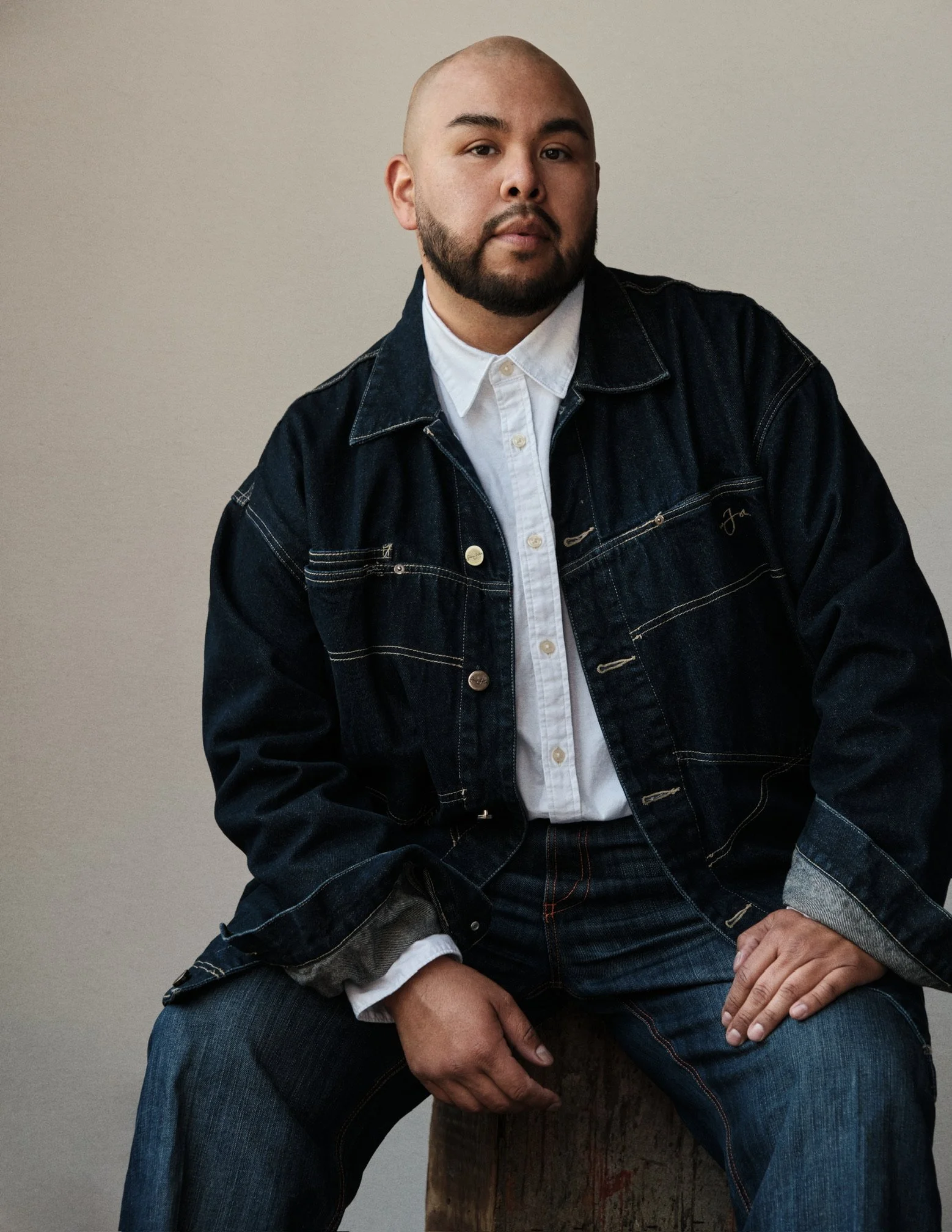 A man with a beard and buzz cut hairstyle, wearing a white shirt and a dark denim jacket, sitting with one hand resting on his knee, against a plain light-colored wall.