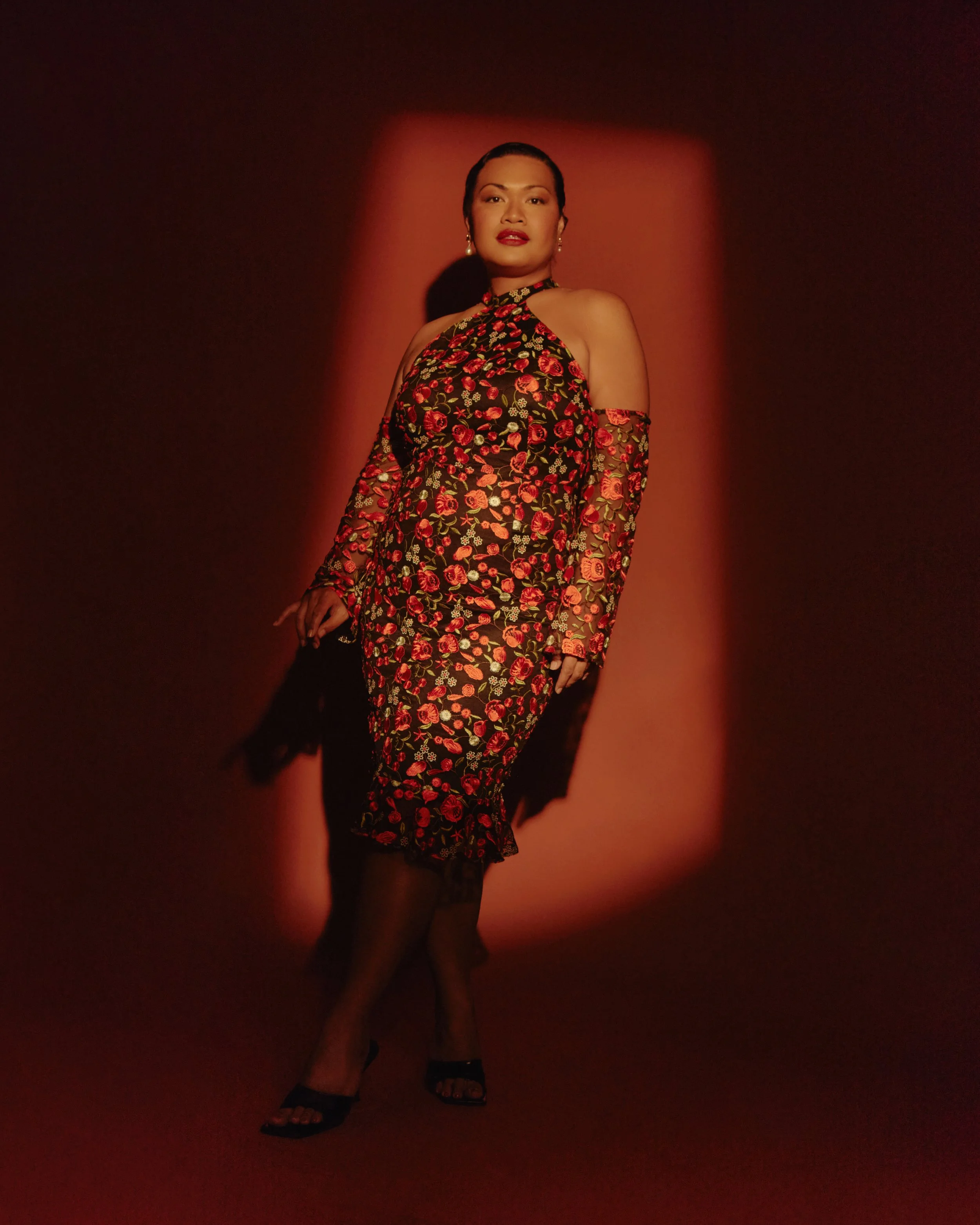 A woman wearing a floral dress with cut-out shoulders stands against a dark background illuminated by a reddish light, with her hands relaxed by her sides and a confident expression on her face.