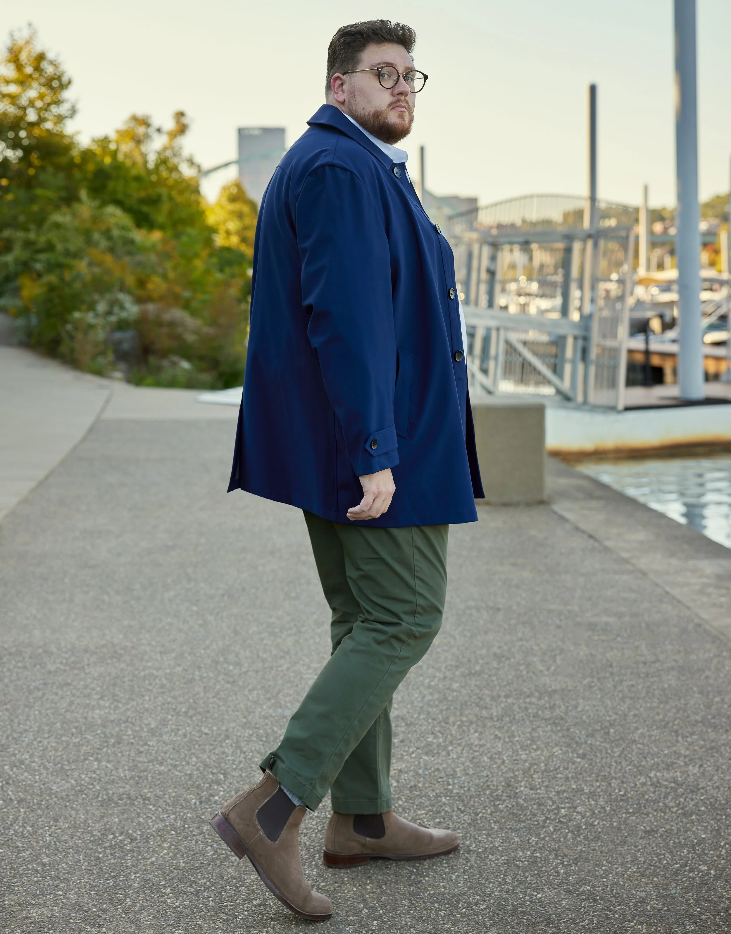 A man wearing glasses, a blue jacket, green pants, and brown boots standing on a sidewalk near a parking lot with boats, trees, and a bridge in the background.