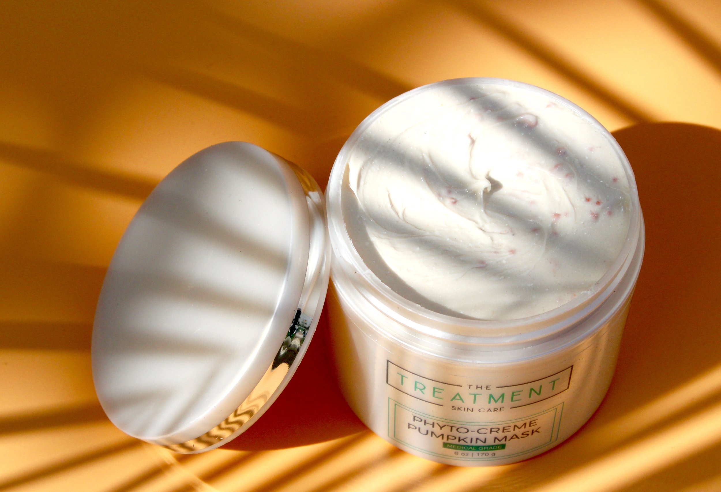 Open jar of skincare treatment cream with spoon on a yellow surface.