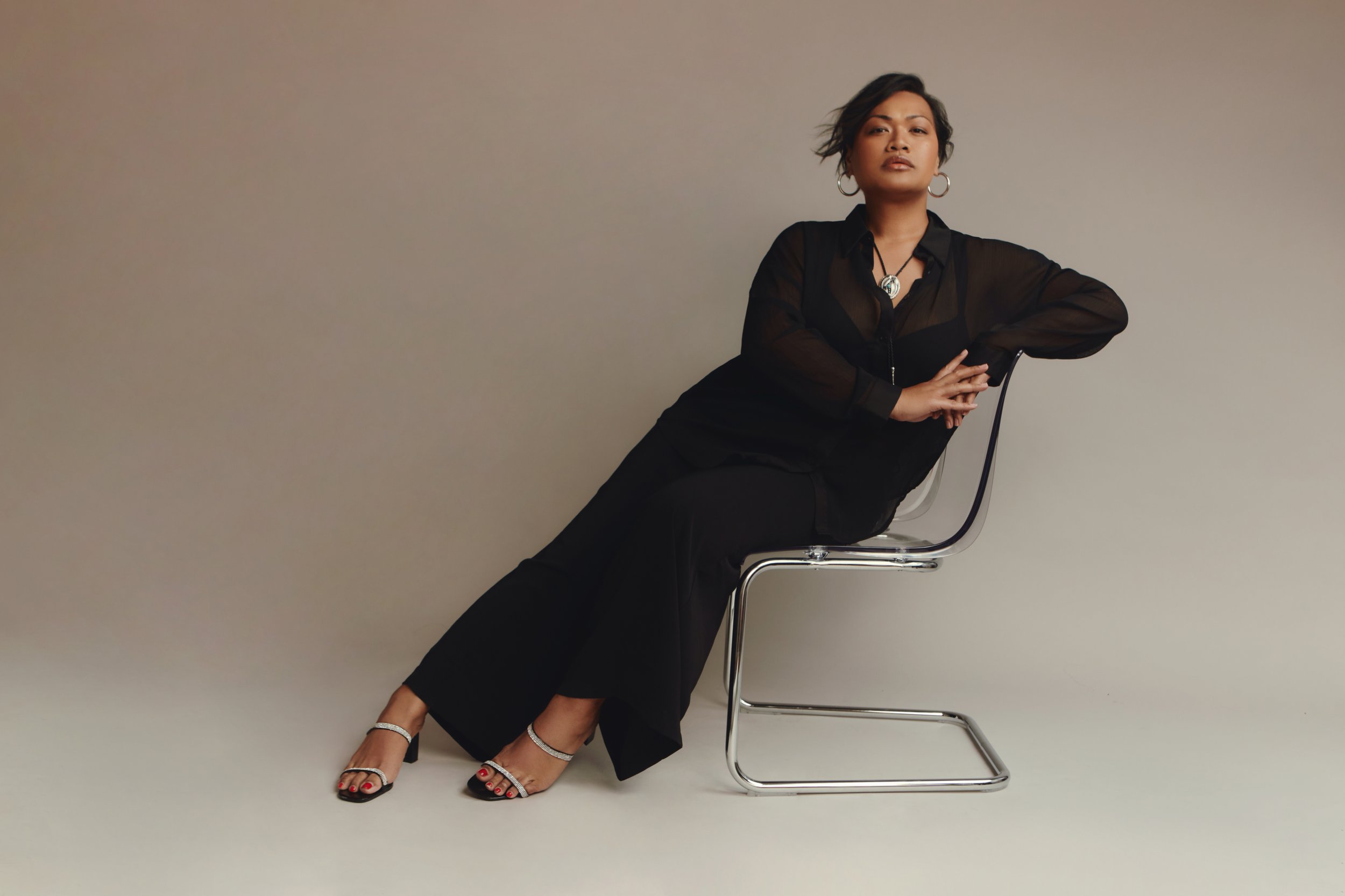 A woman sitting sideways on a modern chair against a plain background, wearing a black sheer top, black wide-leg pants, silver heels, and accessorized with large hoop earrings and a necklace. She has short, styled hair and an expression of confidence.