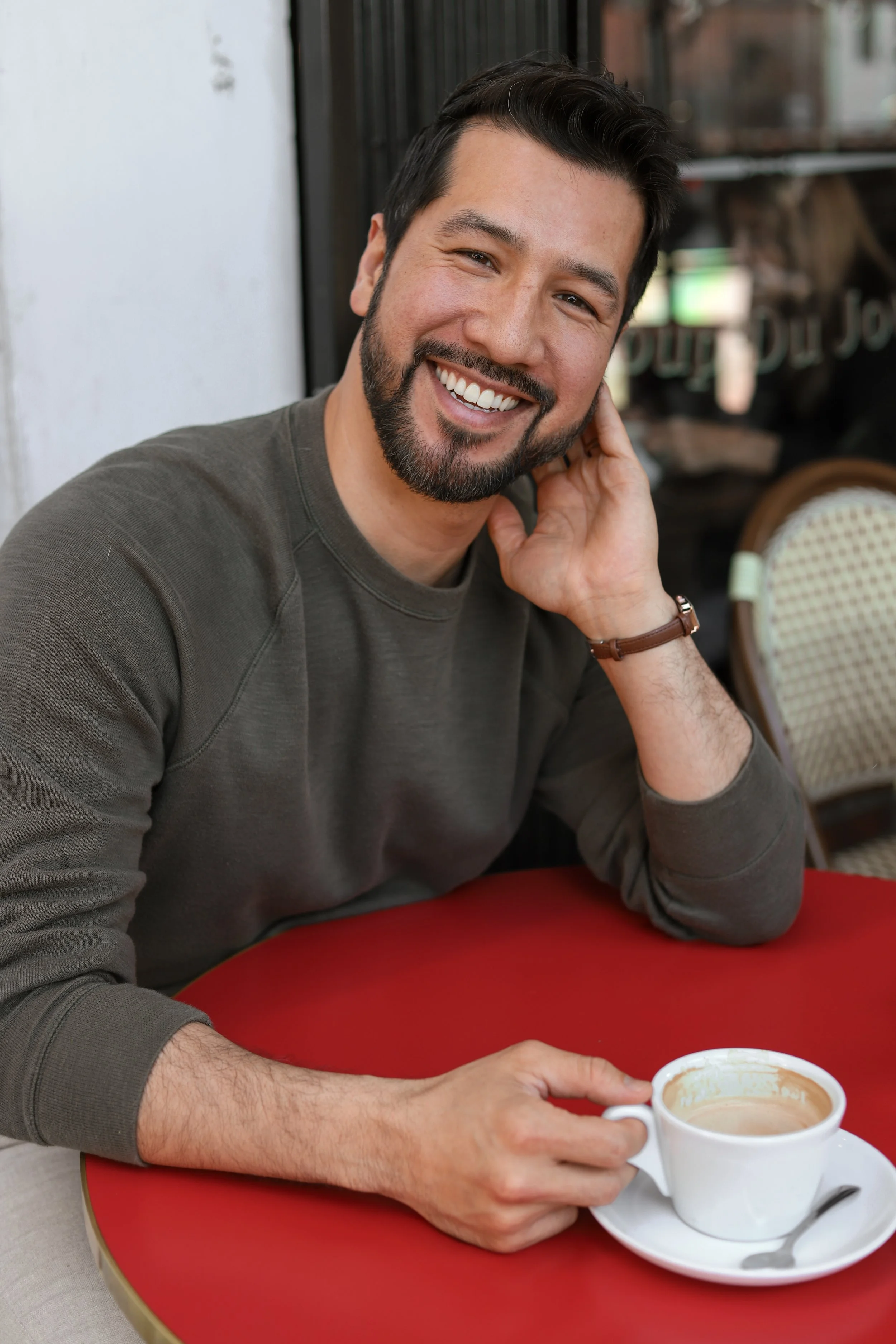 A smiling man with dark hair, beard, and mustache, sitting at a red table in a cafe, holding a cup of coffee with a saucer and a spoon. He is wearing a brown long sleeve shirt and a wristwatch.