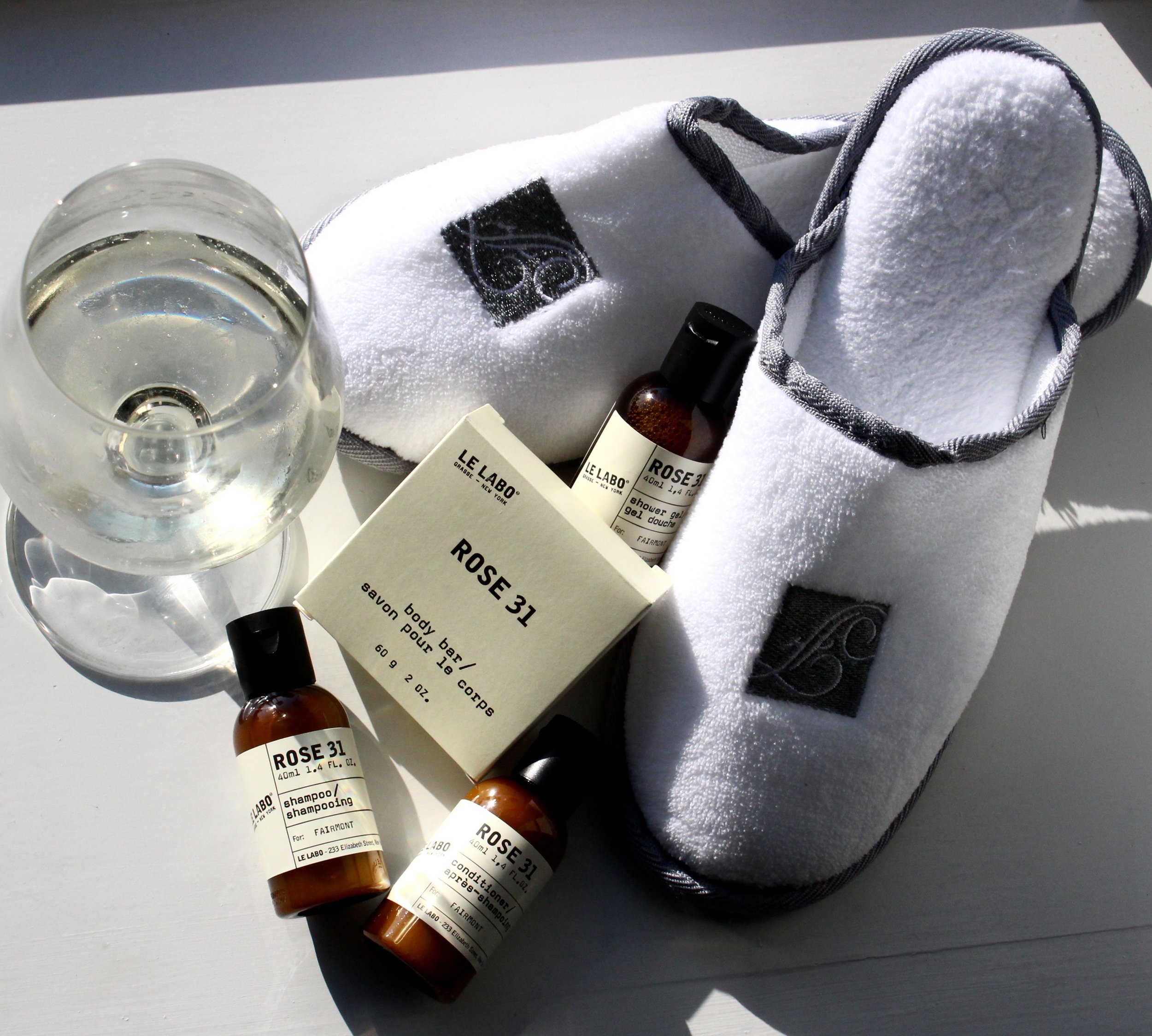 Various skincare and beauty products on a white surface, including a white towel, a glass of water, and bottles of Le Labo Rose 31 bath and body products.