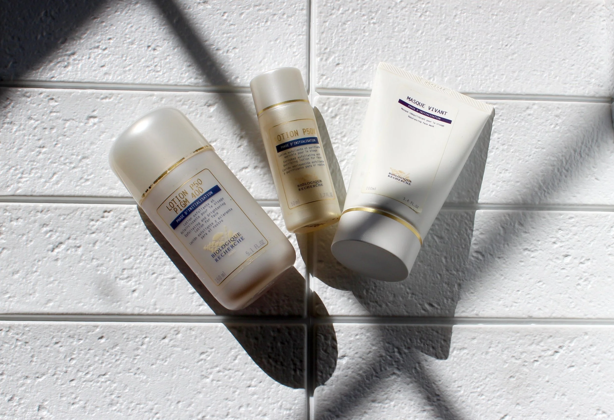 Four bottles of skincare products lying on a white textured surface with shadows cast over them.