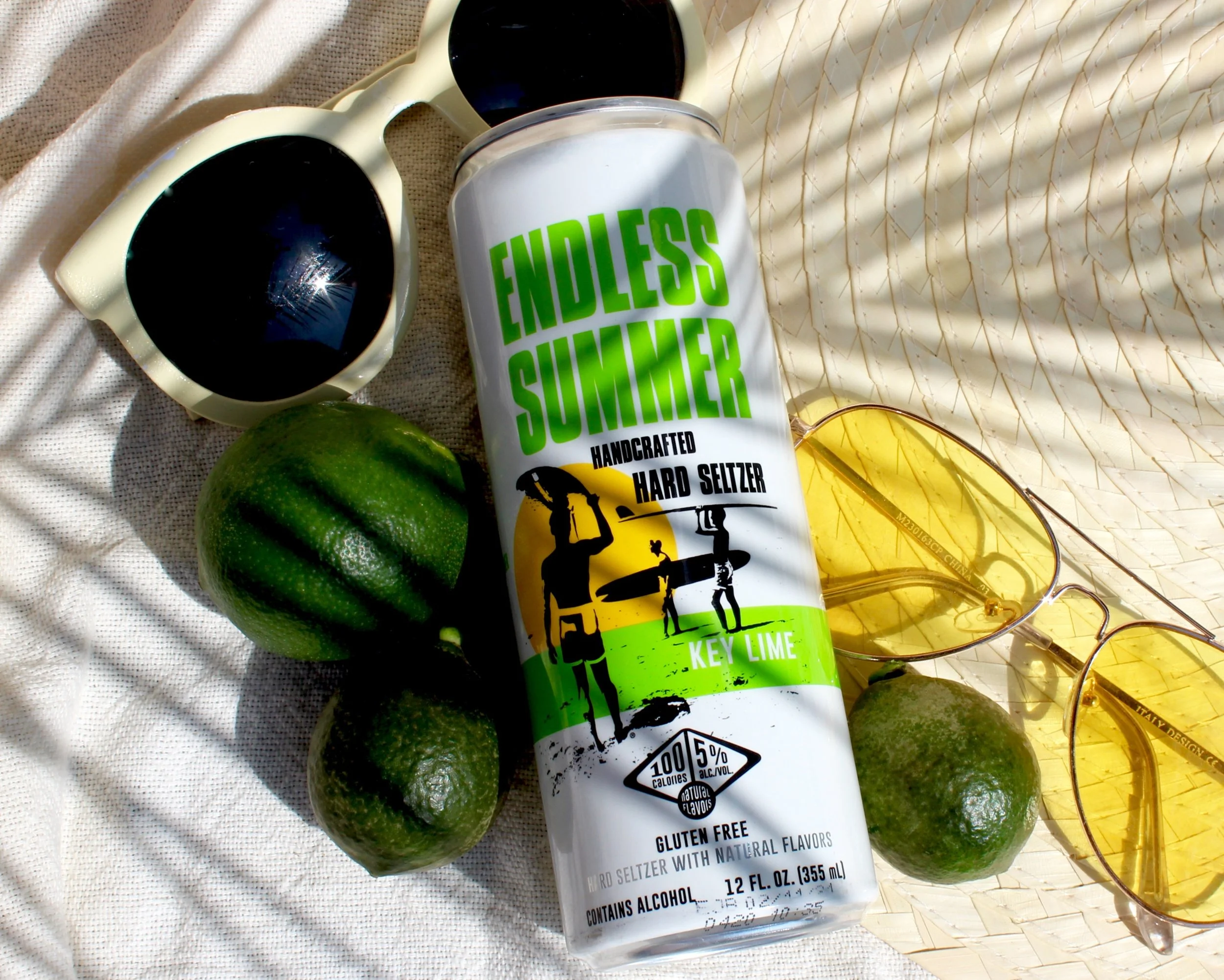A white can of handcrafted hard seltzer with lime flavor, surrounded by three ripe limes, sunglasses, and a pair of yellow earrings, on a beige fabric background with sunlight and shadows.